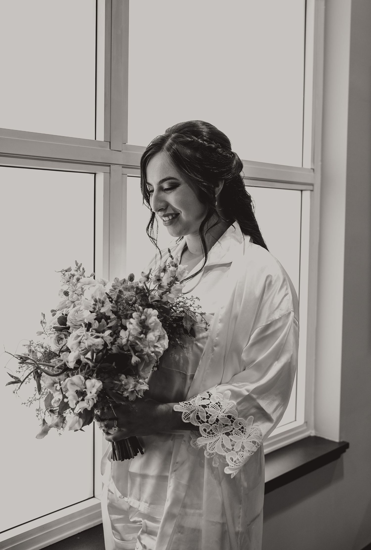 Bride in white robe smiling while holding a floral bouquet near a window, black and white wedding photo.