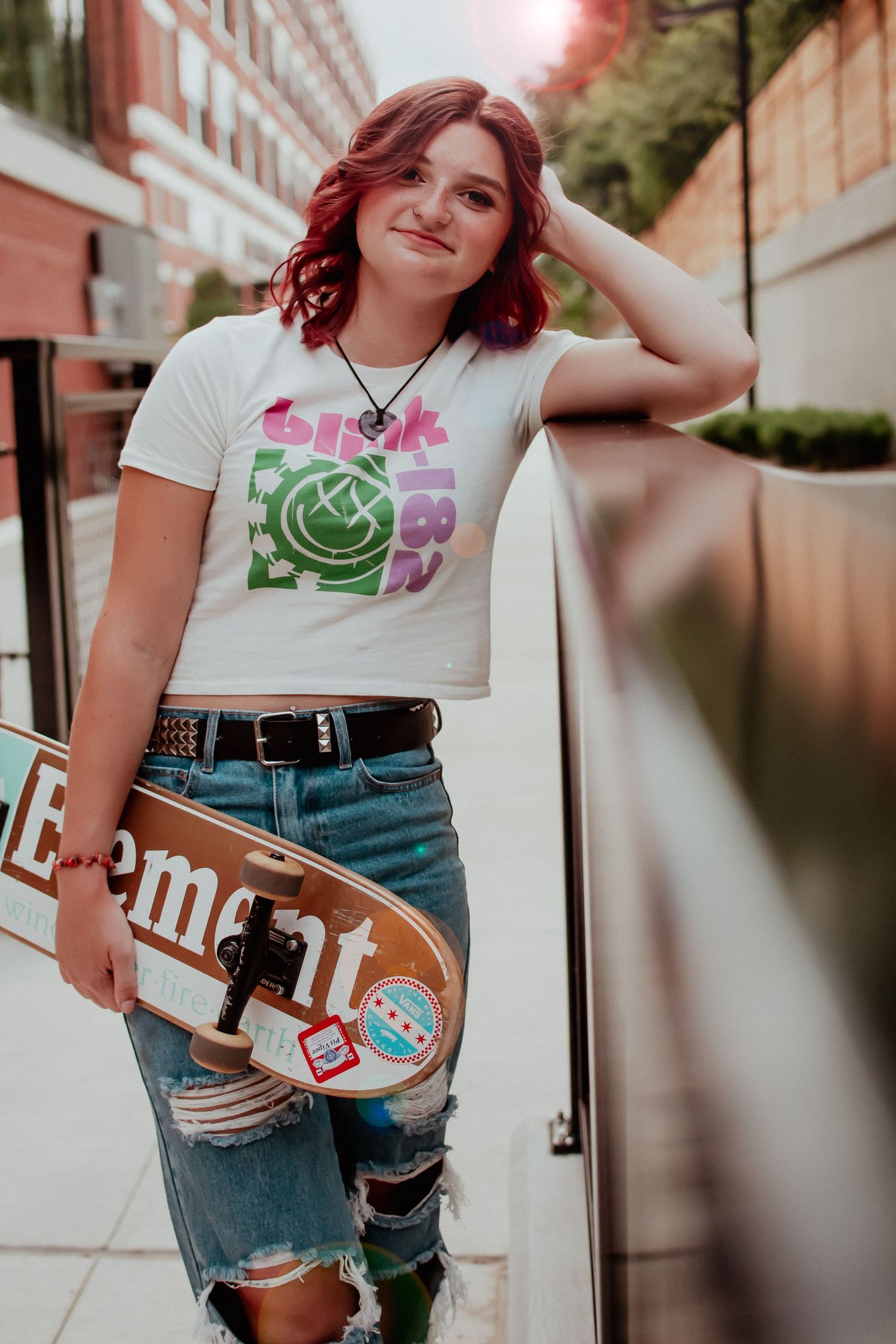 Urban skater style with graphic tee and ripped jeans holding skateboard against brick wall.