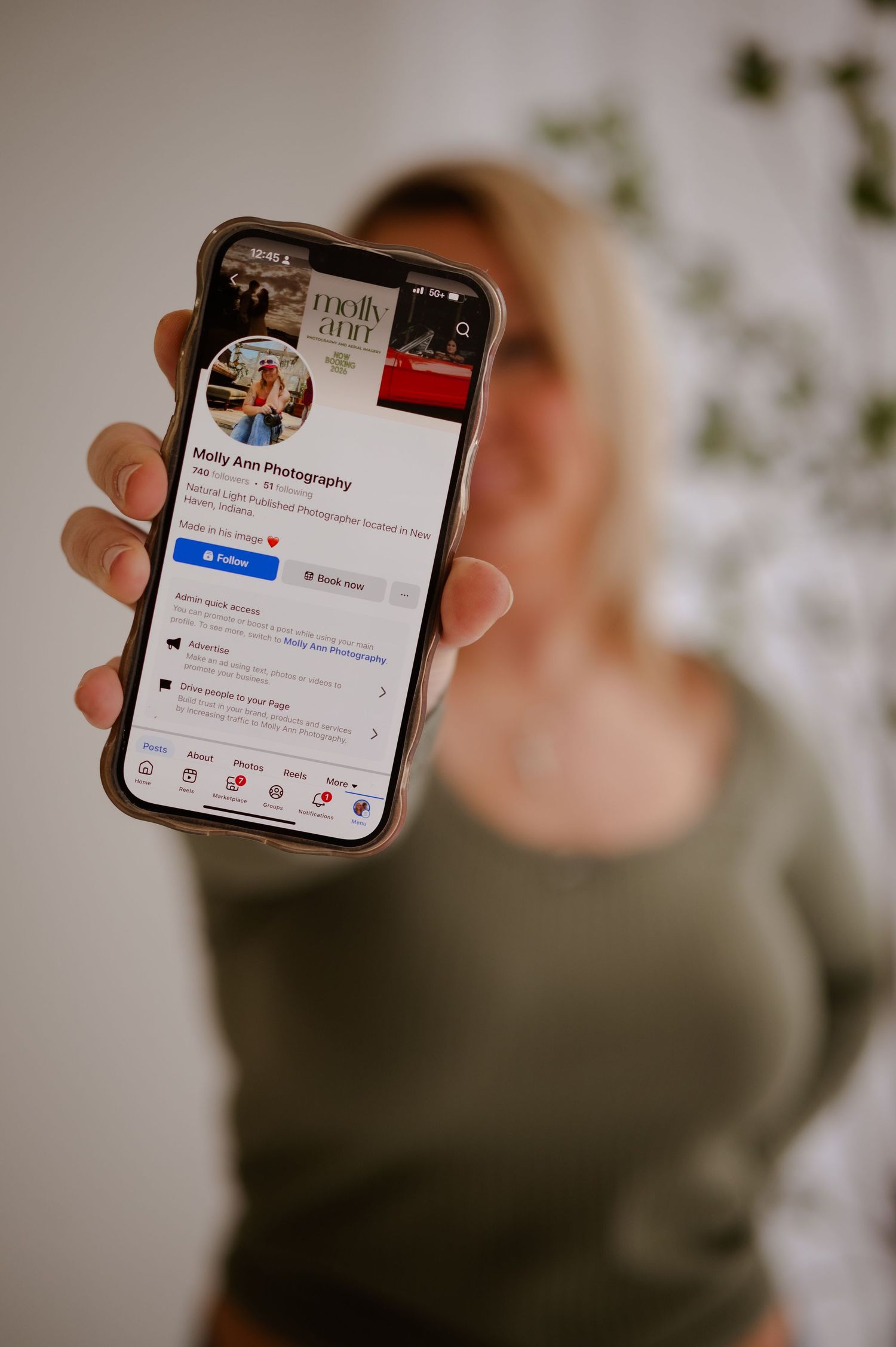 Blonde woman holding smartphone displaying Molly Ann Photography Facebook page profile in portrait orientation.