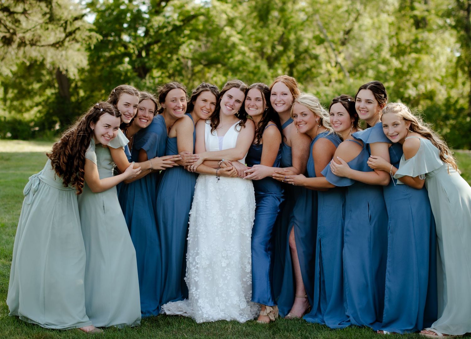 Bridesmaids in blue dresses huddle around bride for fun group photo on lawn.