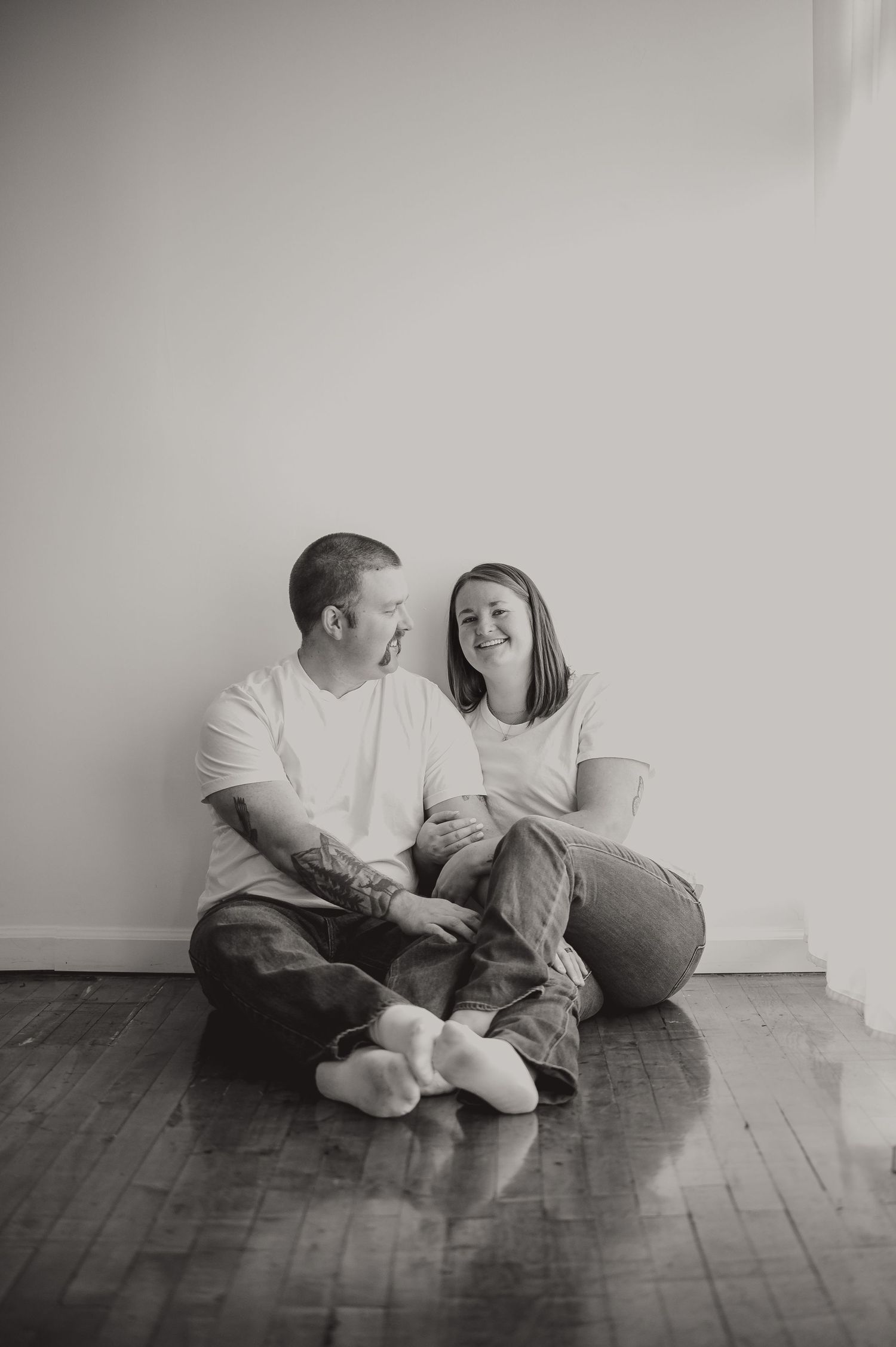 A casual couple sitting on hardwood floors sharing a lighthearted moment in black and white.