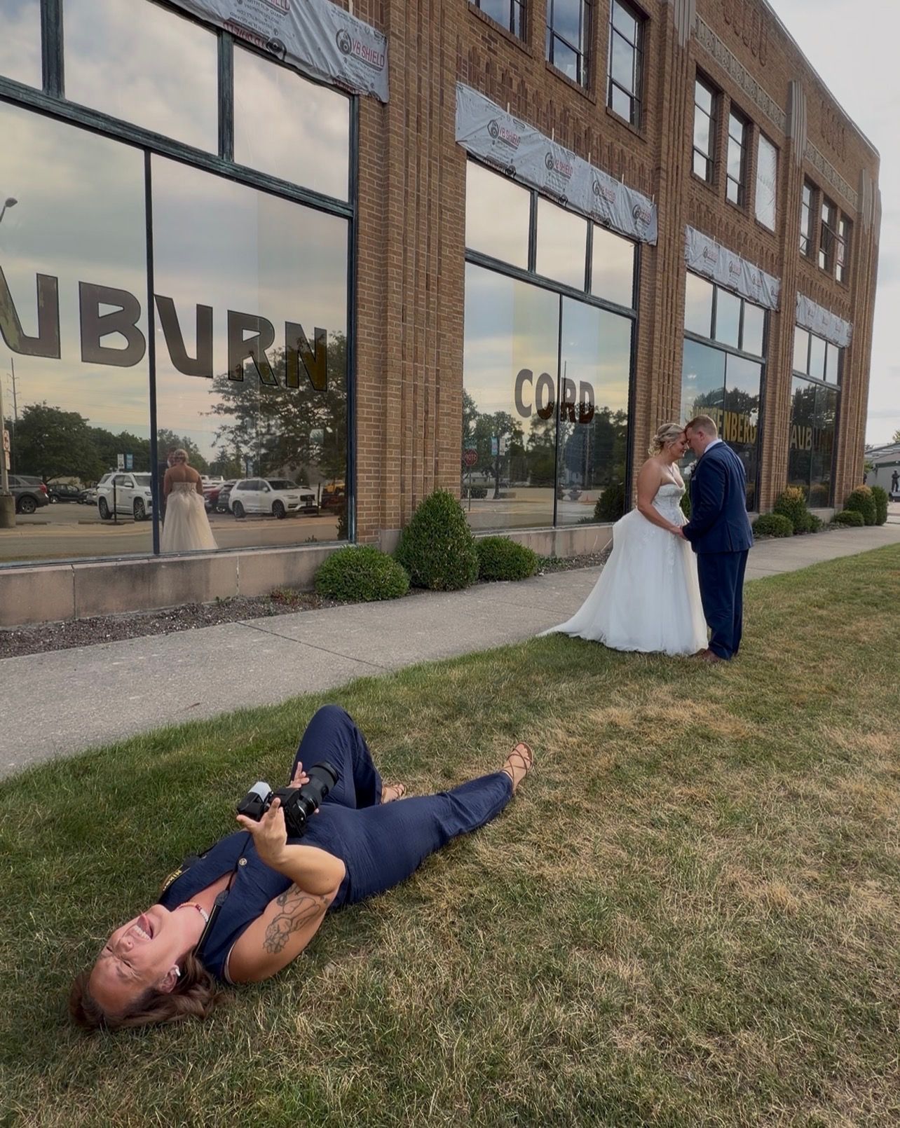 Wedding photographer lies on grass to capture bride and groom posing outside Auburn Cord brick building.