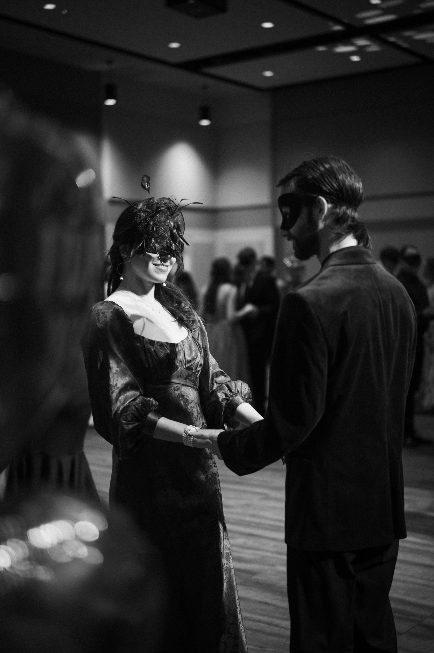 A couple dances together at a masked ball, wearing elegant formal attire in a dimly lit ballroom.