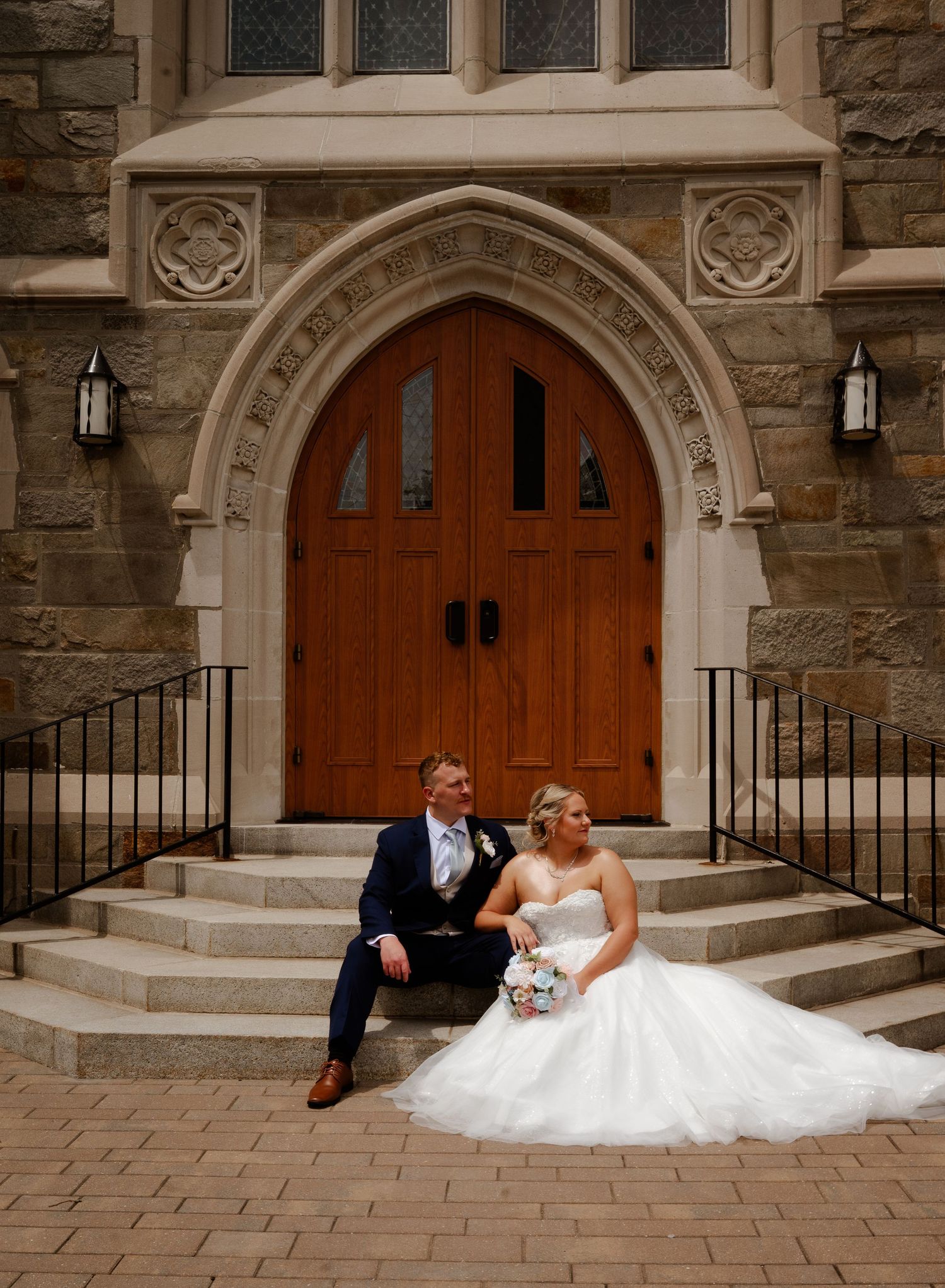 Bride and groom sitting on stone church steps, bride in white gown holding bouquet, groom in navy suit, ornate wooden doors behind.