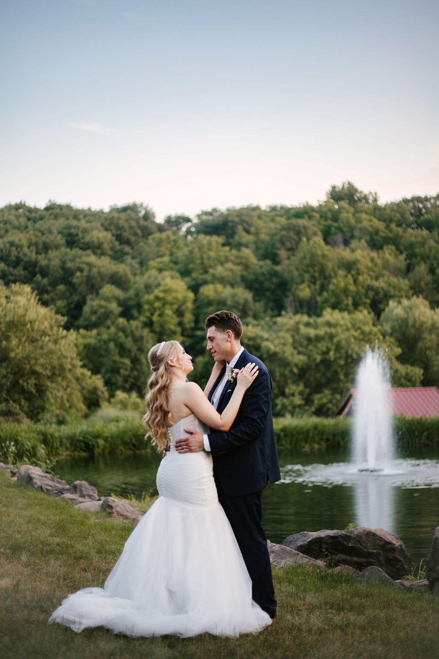 Romantic couple poses by lakeside fountain with lush green landscape in background.