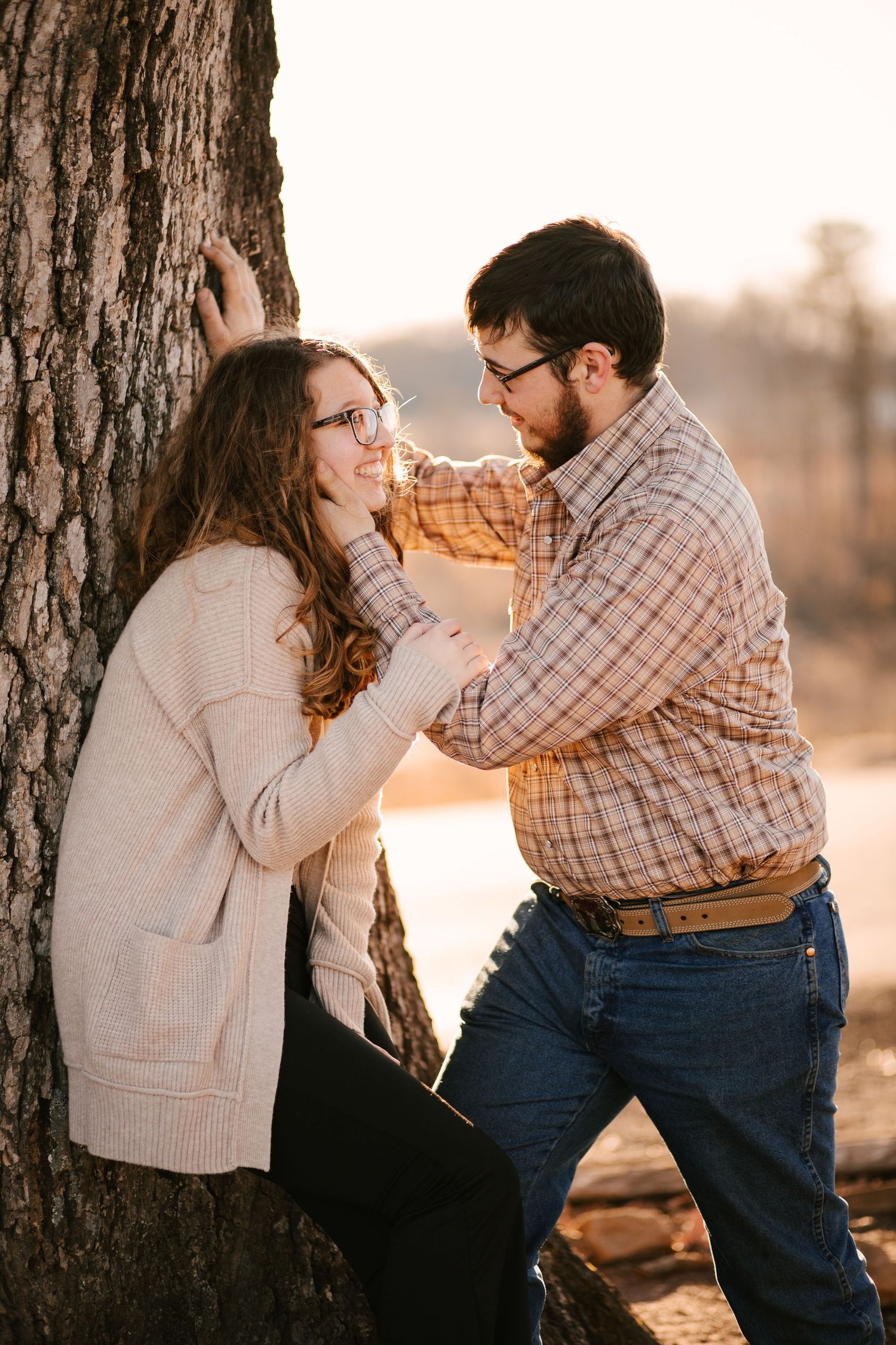 Couple playfully interacts by a tree during sunset in matching casual outfits.