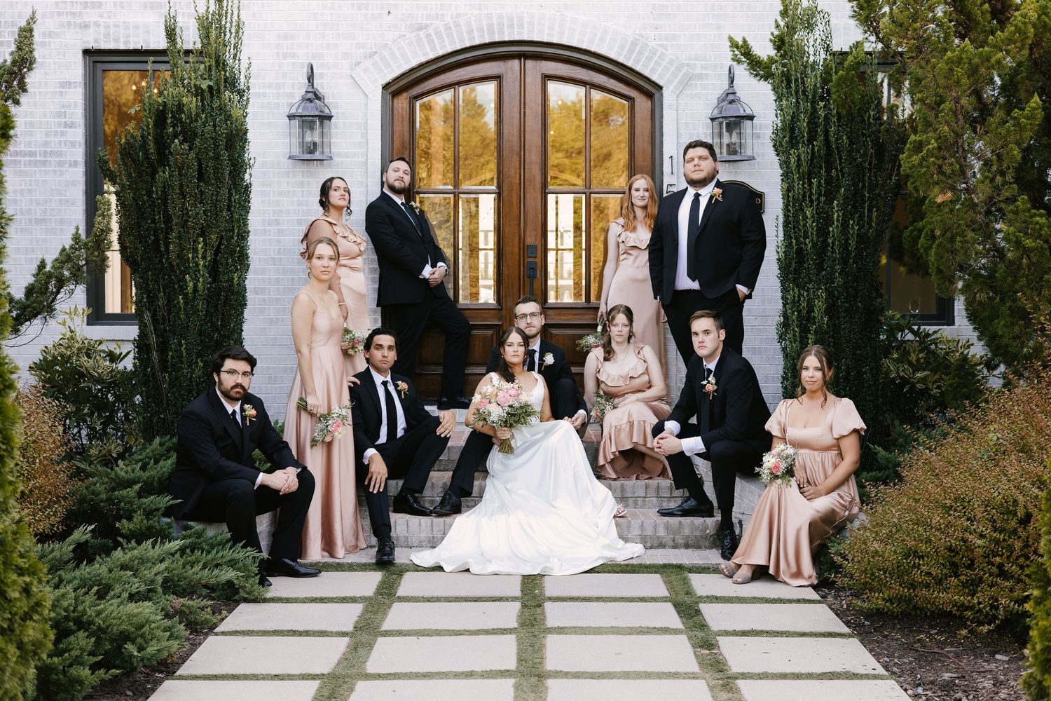 Elegant wedding party poses outside white chapel with cypress trees.