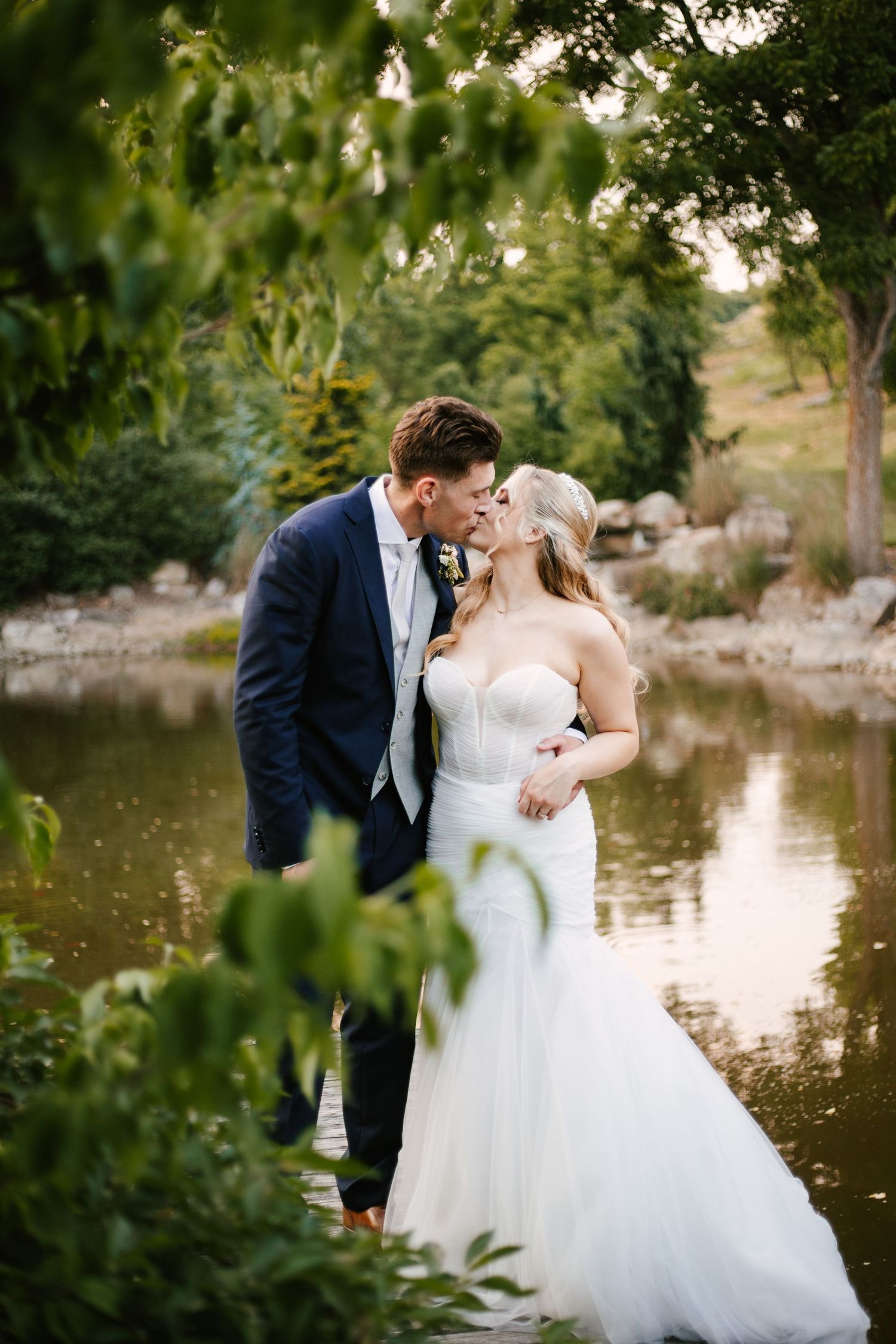 Romantic wedding couple shares intimate moments by a serene pond surrounded by lush greenery at sunset.