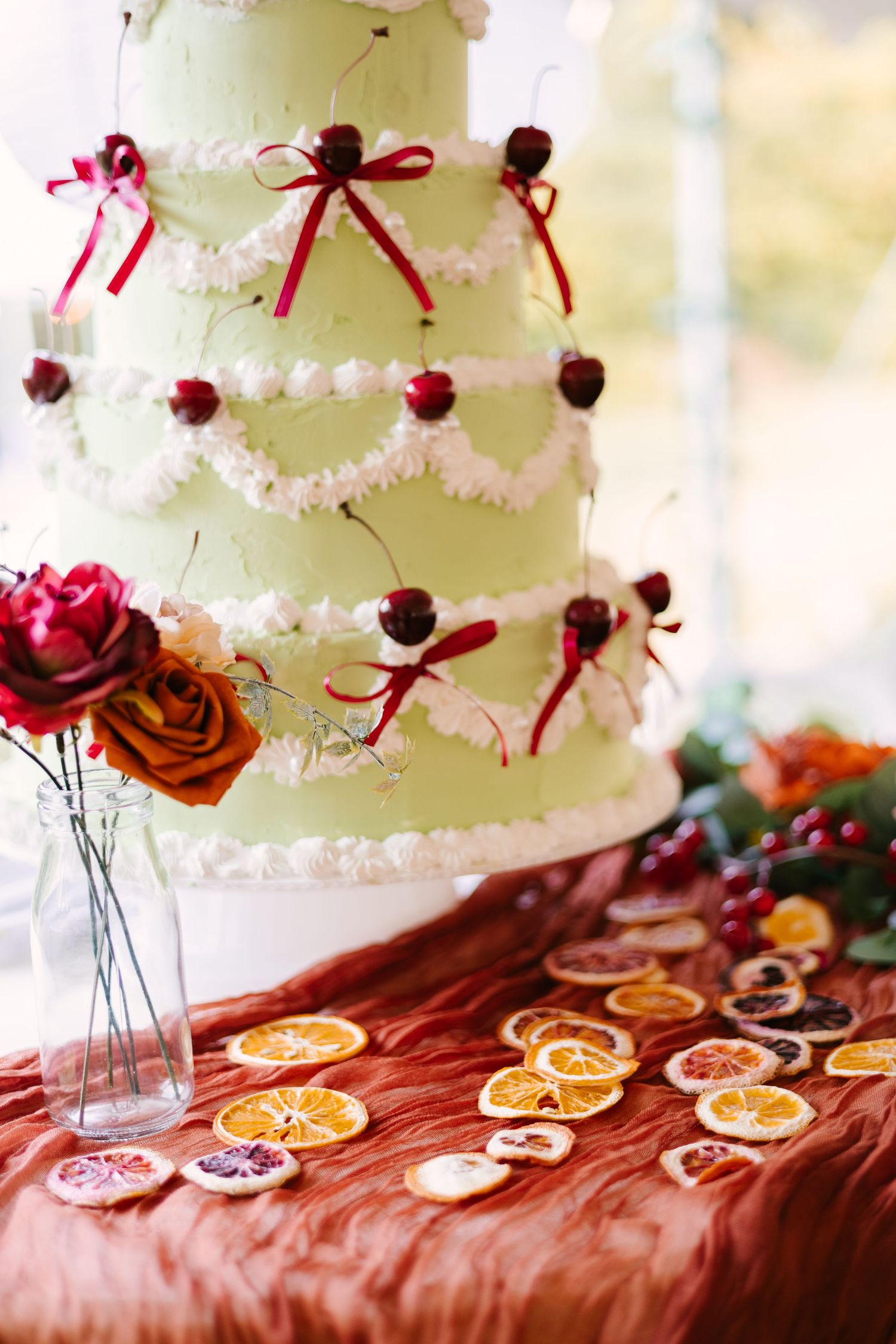 White wedding cake decorated with red ribbons and cherries displayed with dried orange slices.