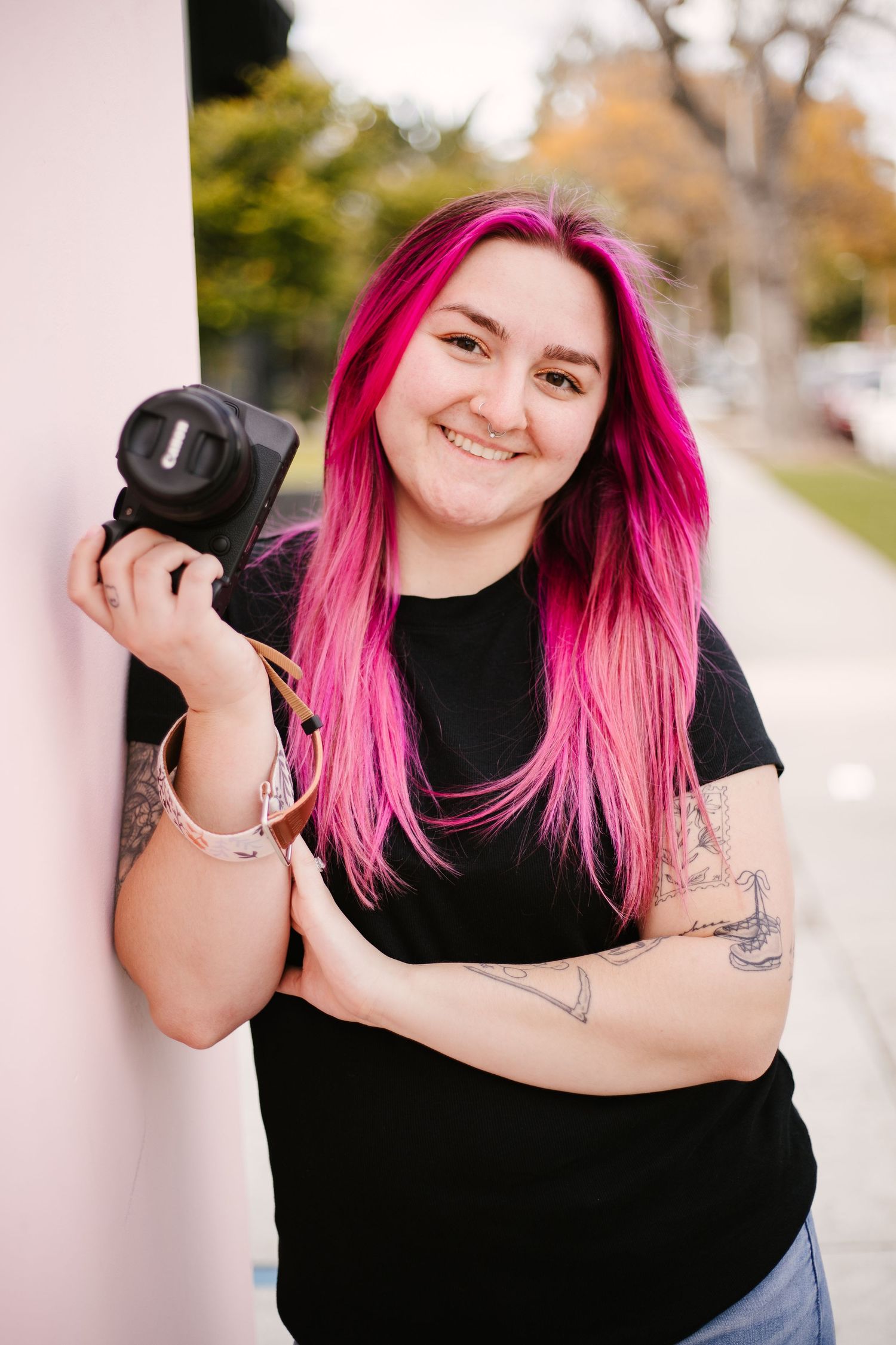 Professional photographer with vibrant pink hair poses with camera against white backdrop.
