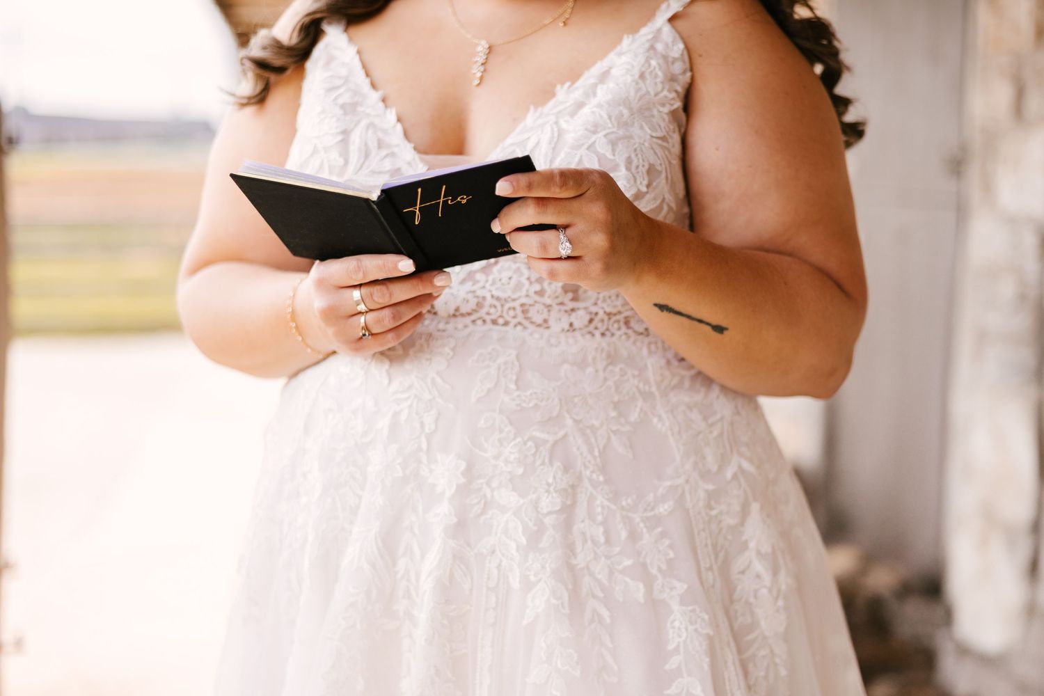 A bride in a lacy white wedding dress holds a leather vow book while standing outdoors.
