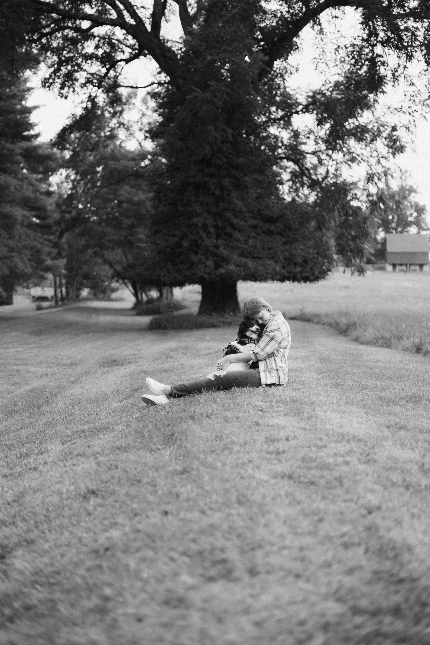 A person relaxes on a grassy hill under large trees in a black and white landscape.