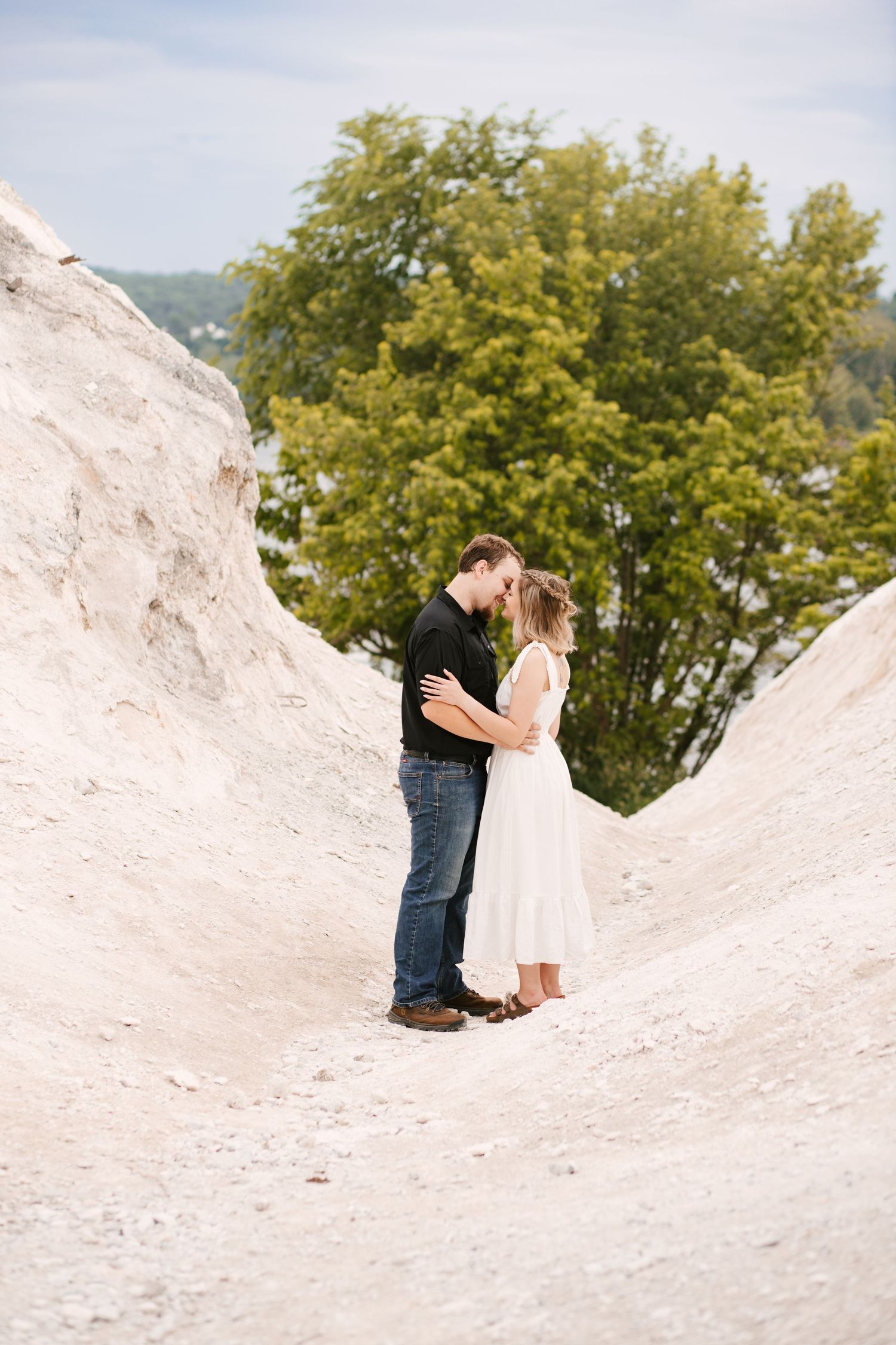 Romantic couple embraces on snowy white terrain with green trees in background.