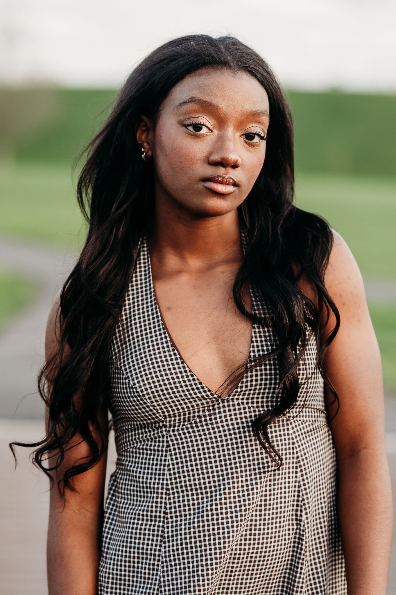 A girl in a black and white gingham sleeveless dress poses outdoors with long flowing dark hair against a blurred background.