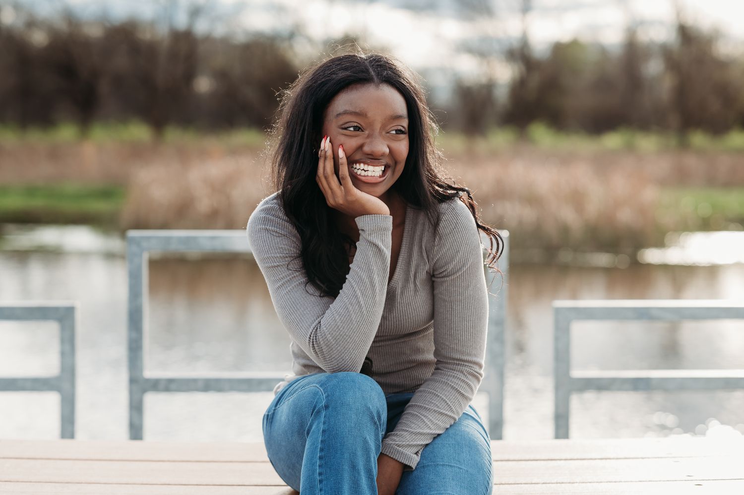 A person in a gray sweater and blue jeans sits casually on a dock by a lake, expressing joy with a bright smile.
