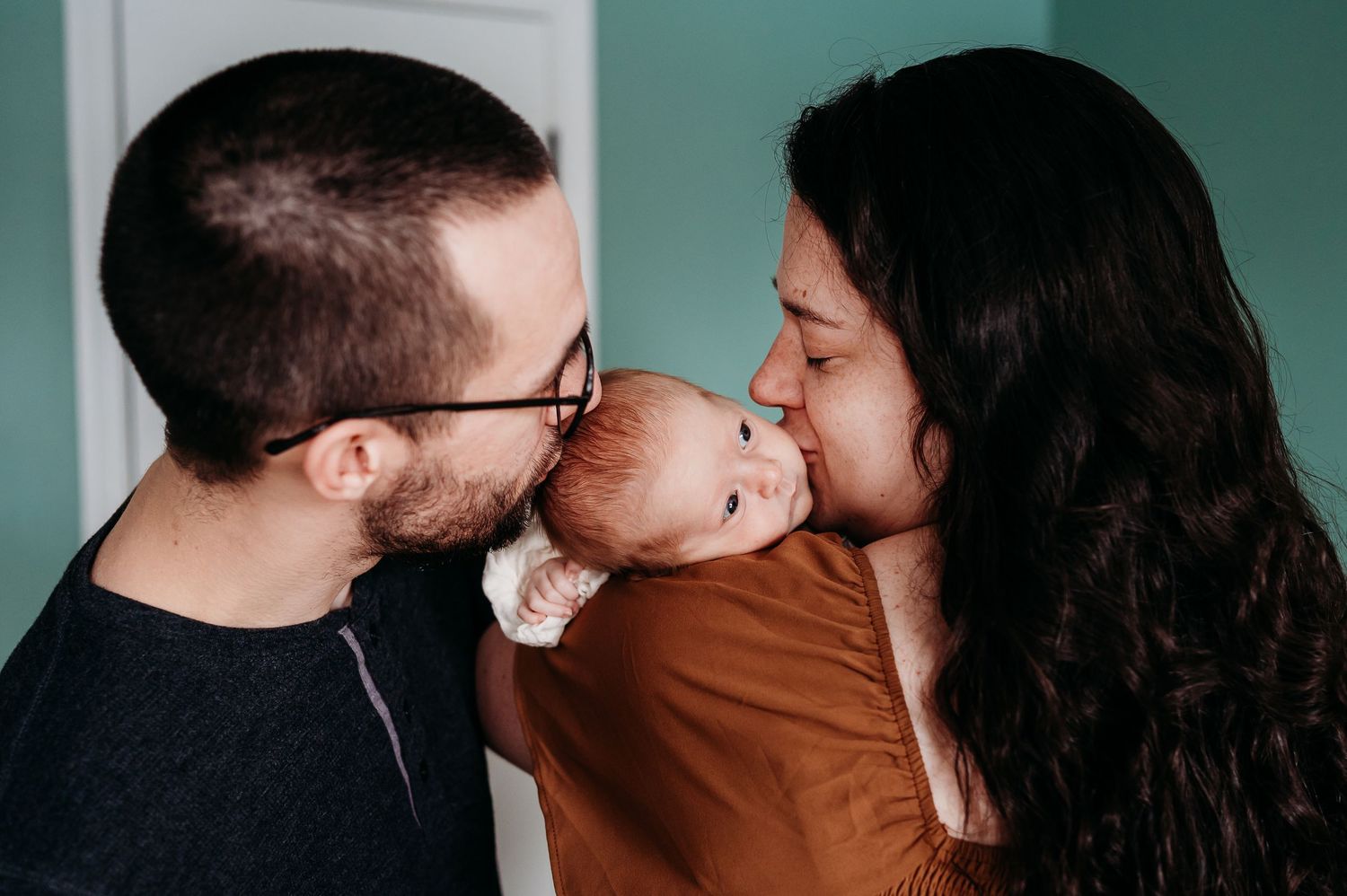 A tender family moment captured against a teal background as Noblesville parents share a kiss with their baby  girl.