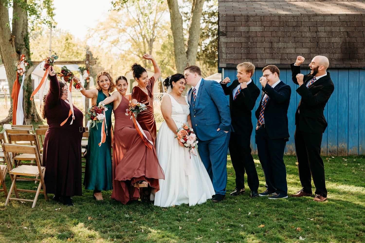 Wedding party in formal attire poses together outdoors with bridesmaids in burgundy dresses and groomsmen in navy suits.