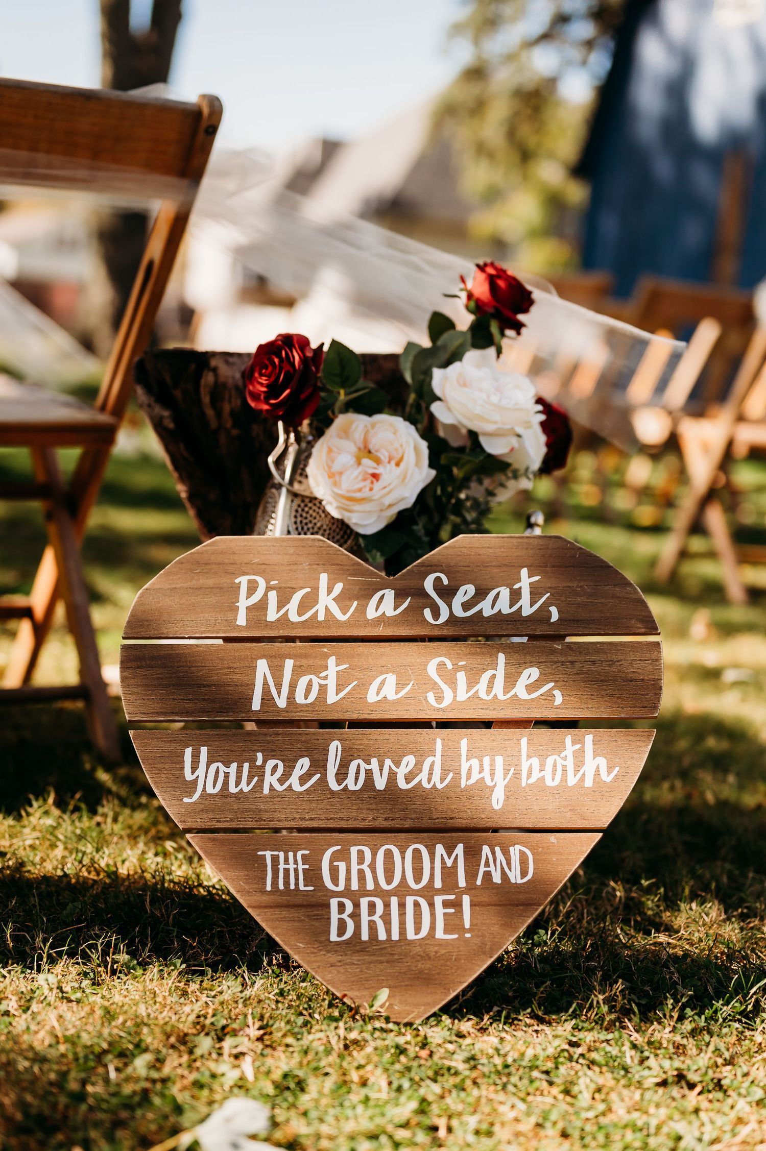 Rustic wooden heart-shaped wedding sign reading 'Pick a Seat, Not a Side, You're Loved by Both' with floral decorations.