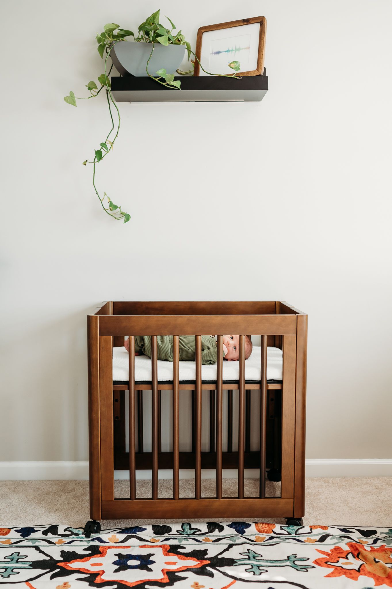 A modern wooden baby crib with baby boy inside beneath a floating shelf with trailing plants and framed artwork.