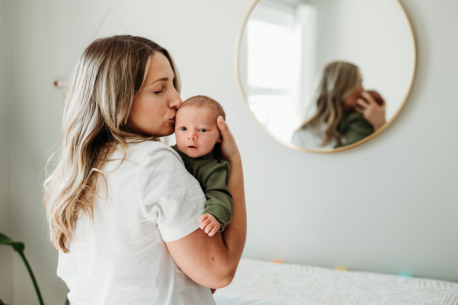 A person in a white shirt holds a newborn baby while standing next to a round mirror on a wall.