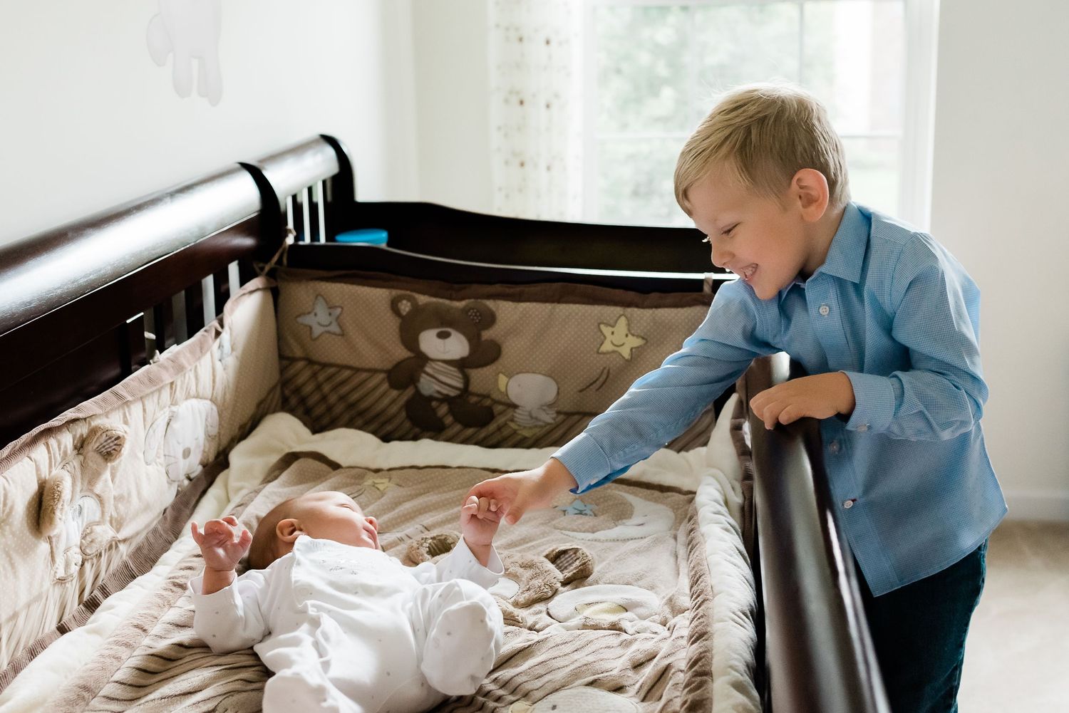 Young child in blue shirt standing at a crib looking down at a baby lying on beige bedding during newborn photoshoot in Avon, Indiana, home.