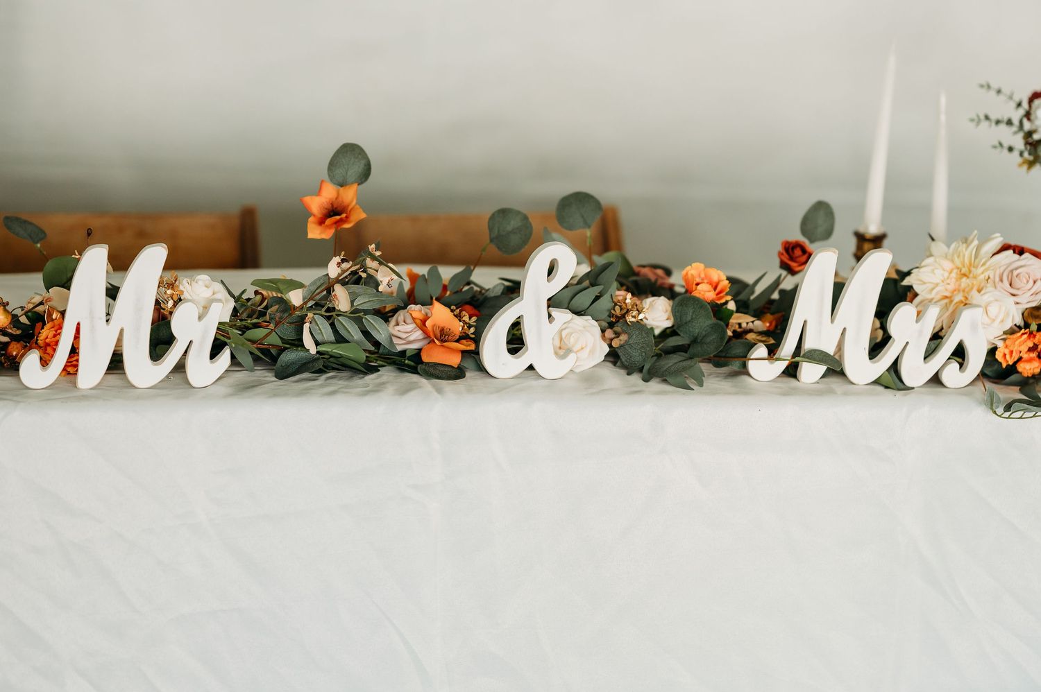 White Mr & Mrs wedding sign decorations displayed on table with orange floral and greenery garland arrangement.