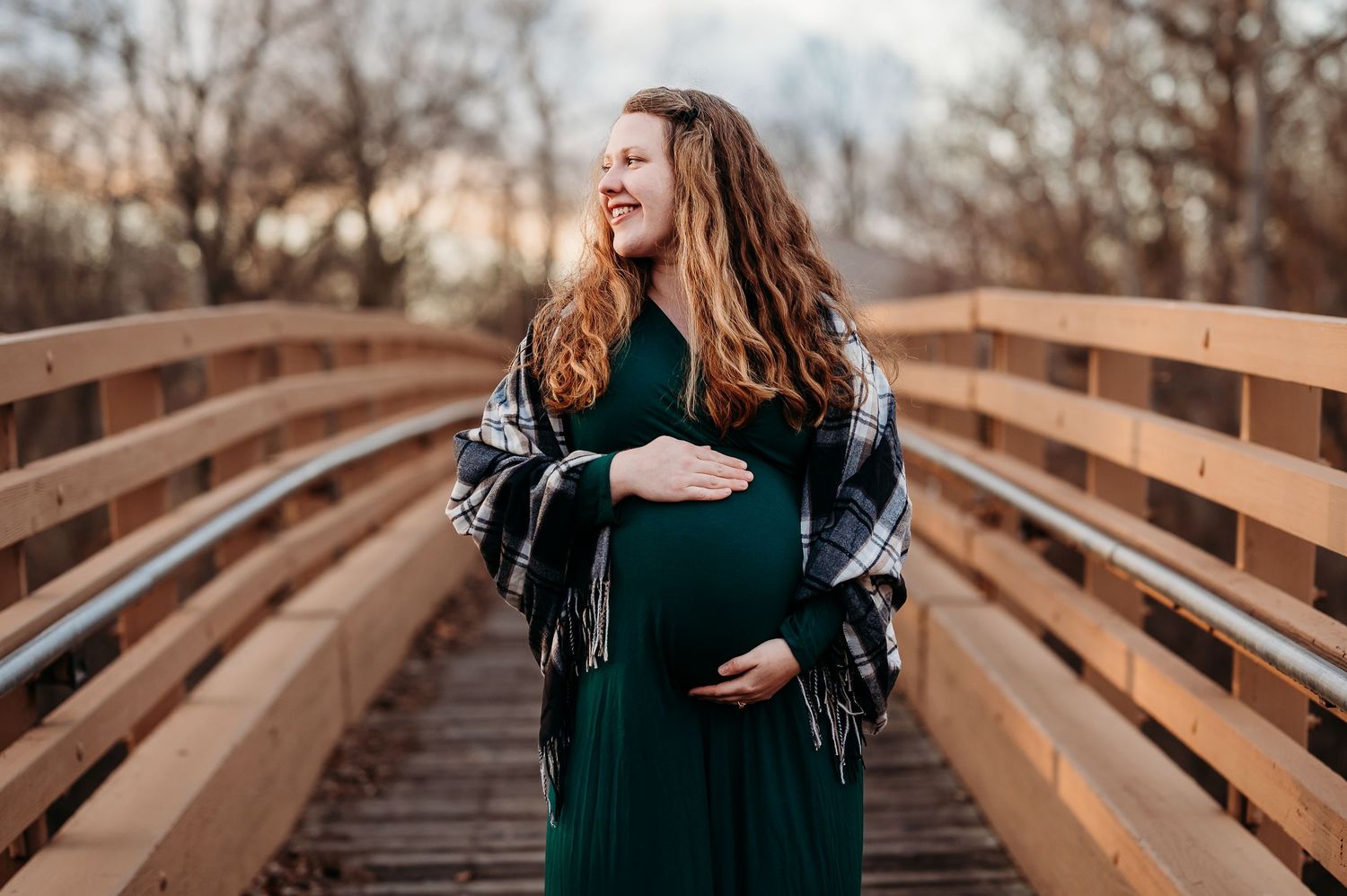 Smiling pregnant woman in green dress and plaid shawl cradling baby bump on wooden bridge outdoors at Plainfield, Indiana, park.