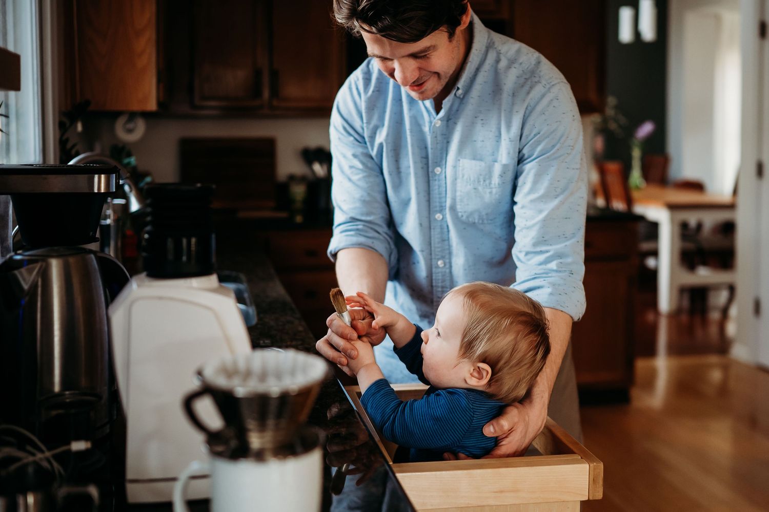 Father smiling while making coffee with his baby toddler in a wooden high chair in a warm, cozy Indianapolis kitchen setting.
