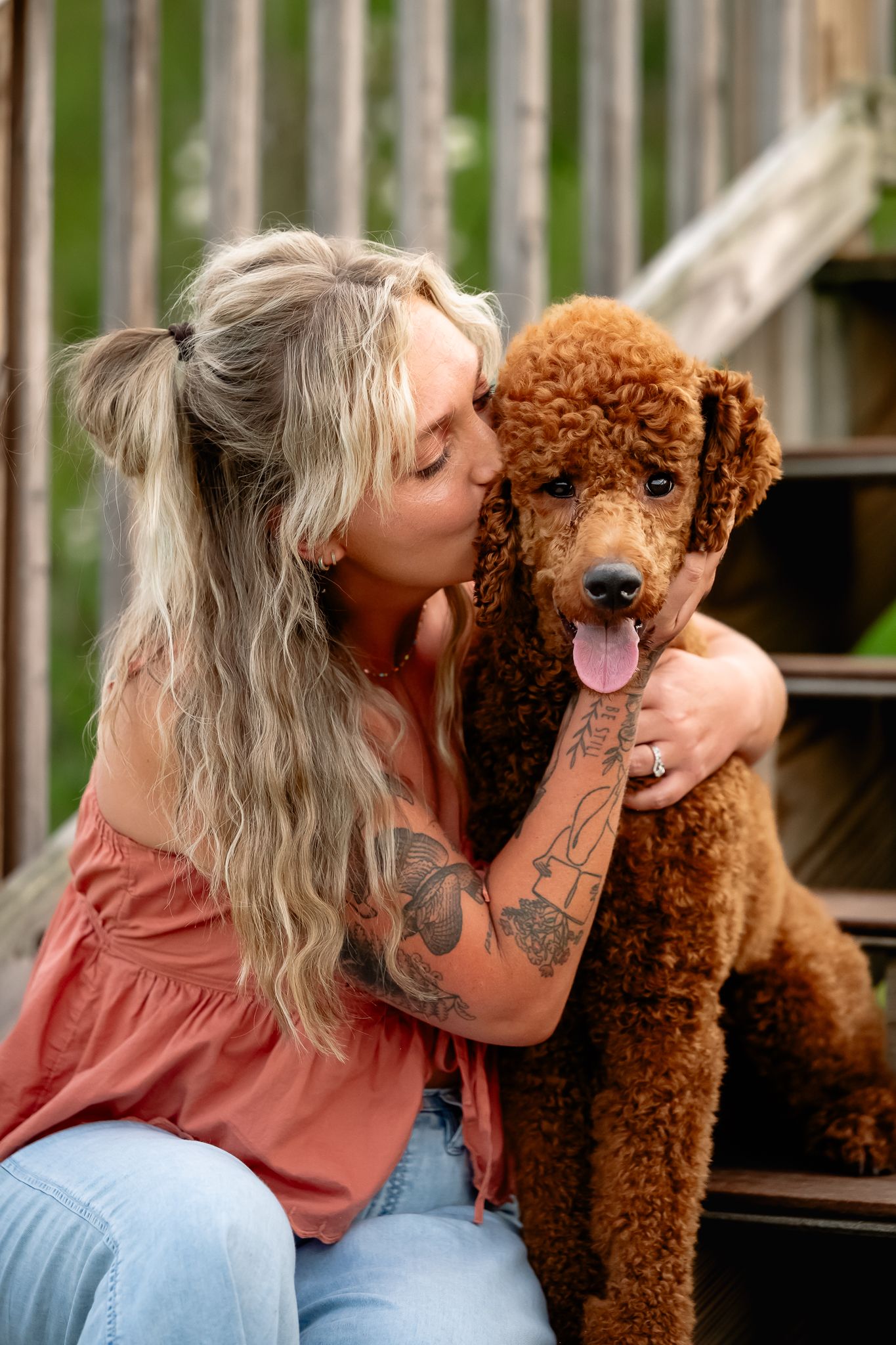 Blonde tattooed woman kissing a fluffy brown standard poodle while sitting on wooden steps outdoors.