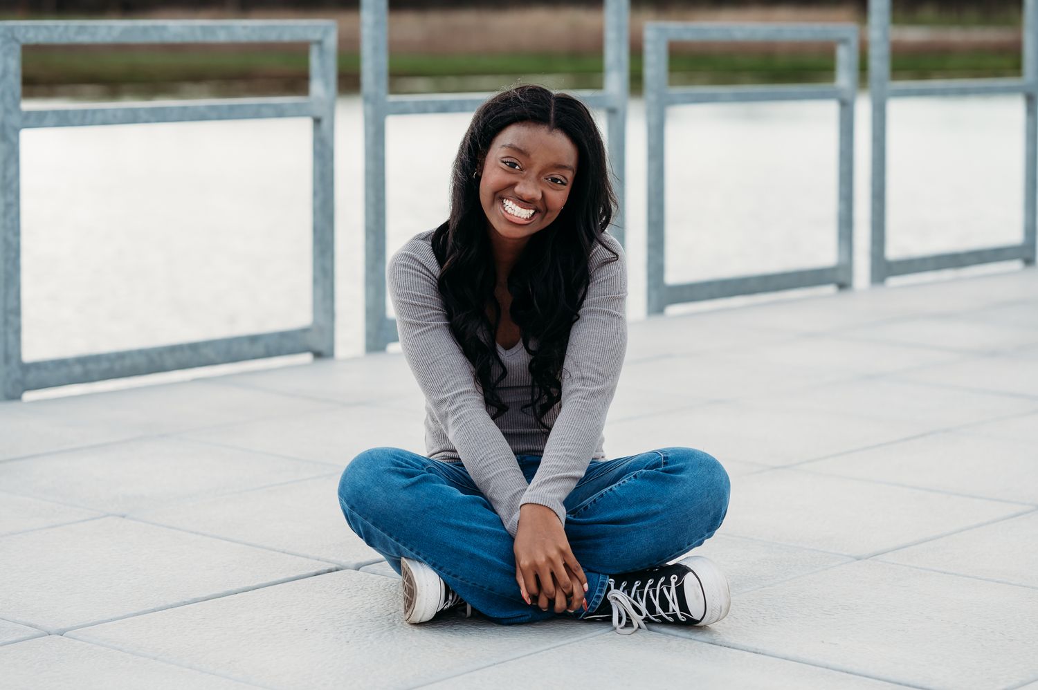 A senior girl in jeans sits on dock while smiling for the camera.