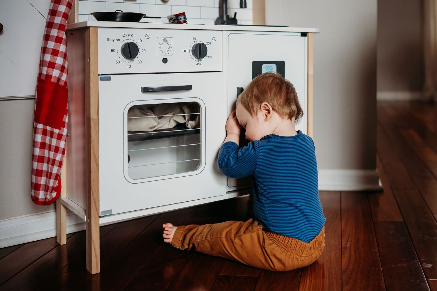 Toddler boy playing with white wooden toy kitchen set on hardwood floor during an Indianapolis in-home photo and video session.