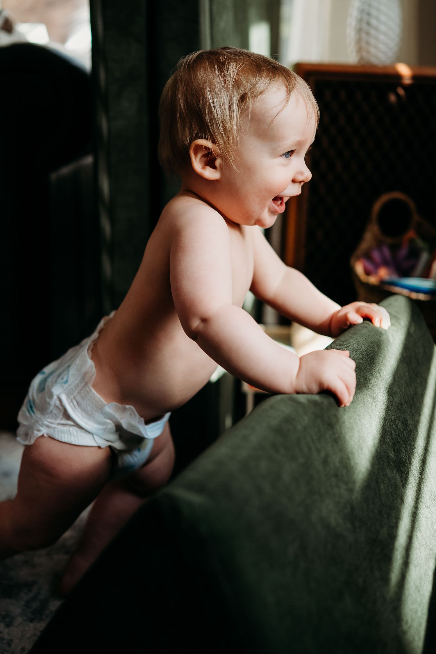 A happy baby in a diaper stands and holds onto a green cushion, learning to walk in a cozy home setting.