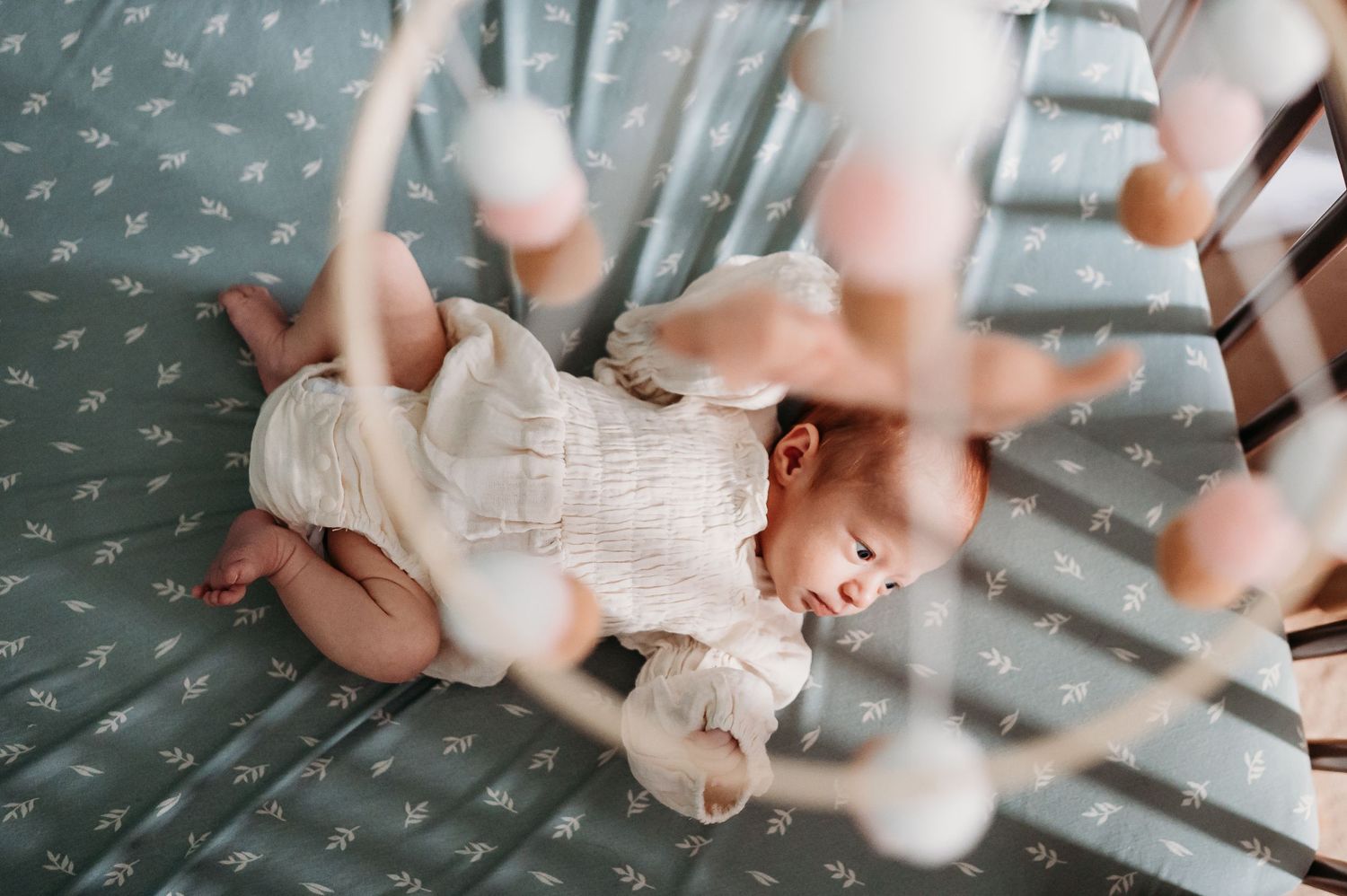 Baby in white outfit lying on polka dot mattress viewed through hanging mobile toys.