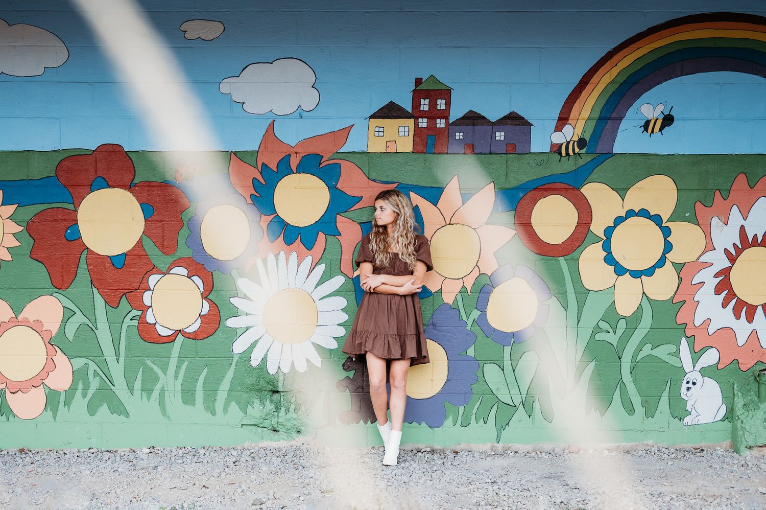 Senior girl stands against colorful mural wall featuring painted flowers, houses and rainbow in whimsical outdoor scene.