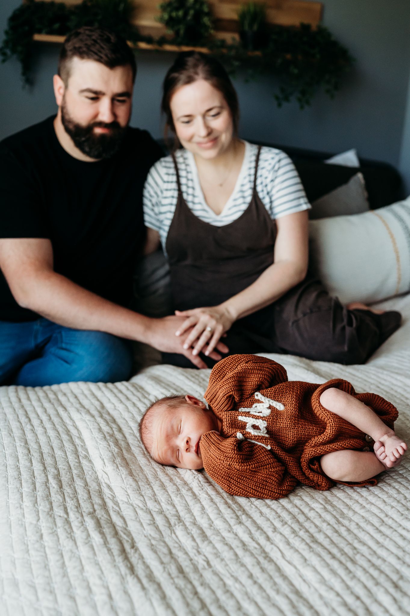 Newborn baby sleeping on white textured bedding wearing an orange knitted sweater while parents watch nearby during Indiana photoshoot.