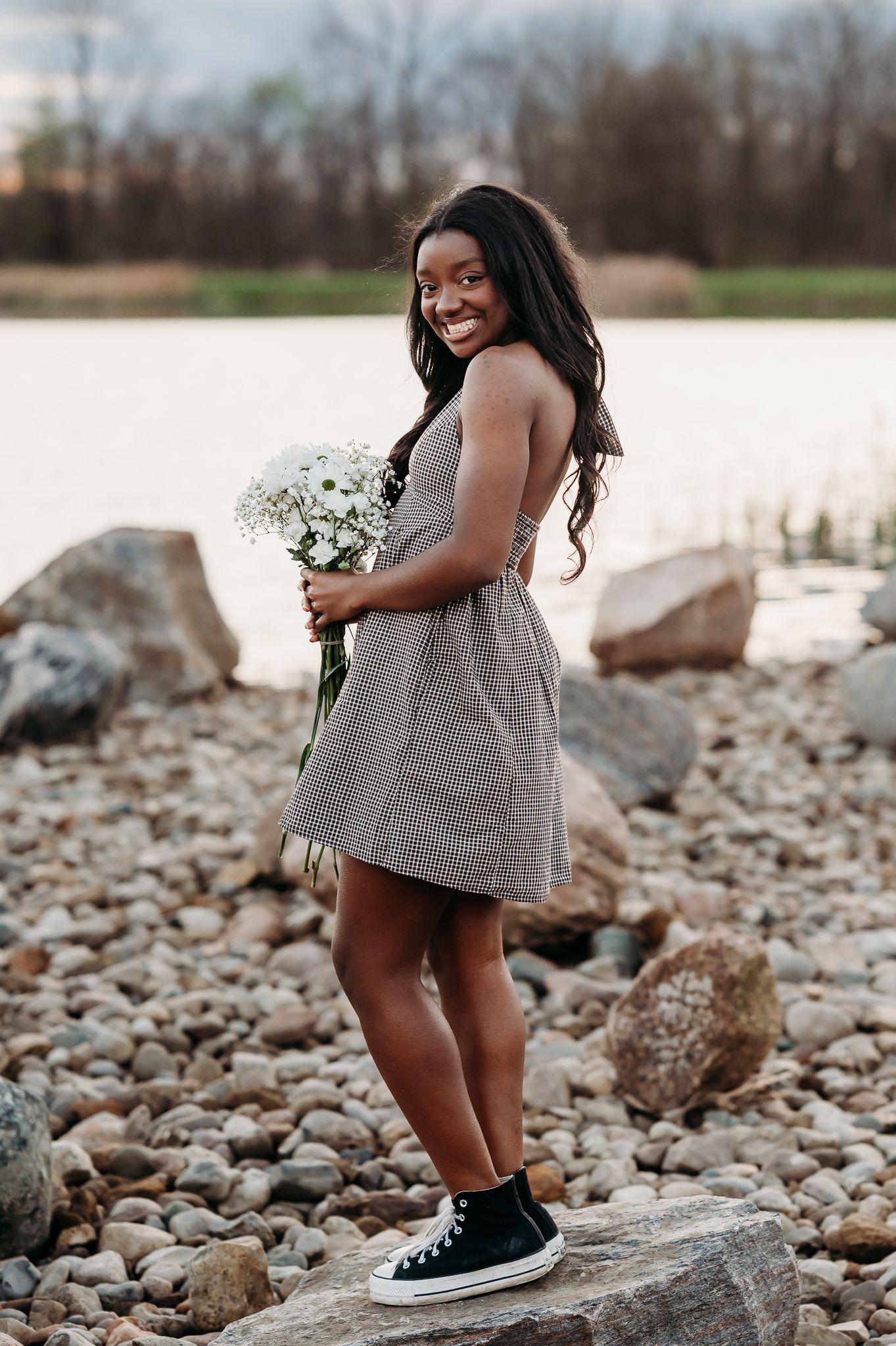 Person in a gray dress holding white flowers poses on rocky shoreline while wearing black sneakers.