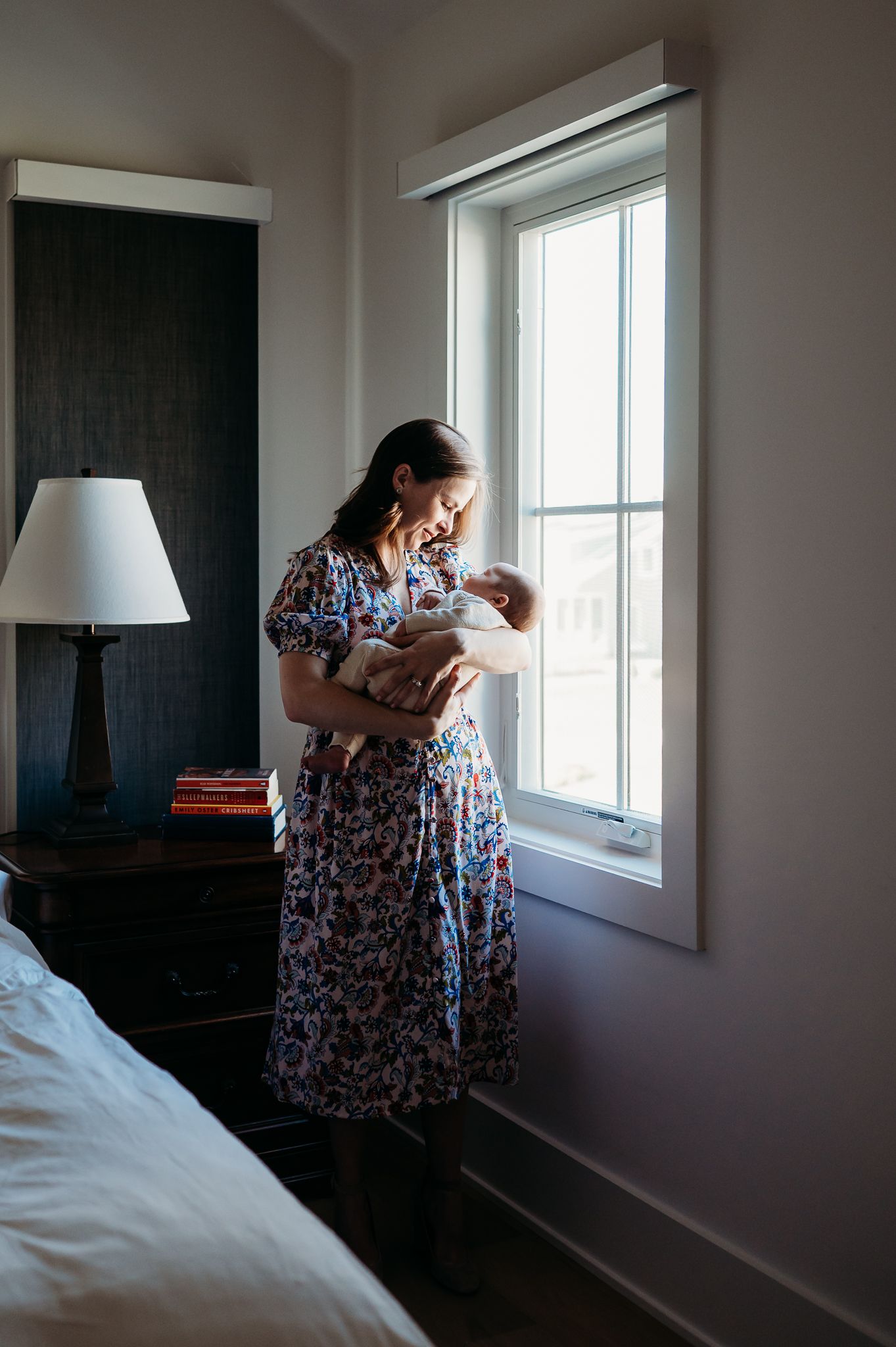 A Westfield woman in a floral dress stands by a bright window while holding a baby in a sunlit bedroom.
