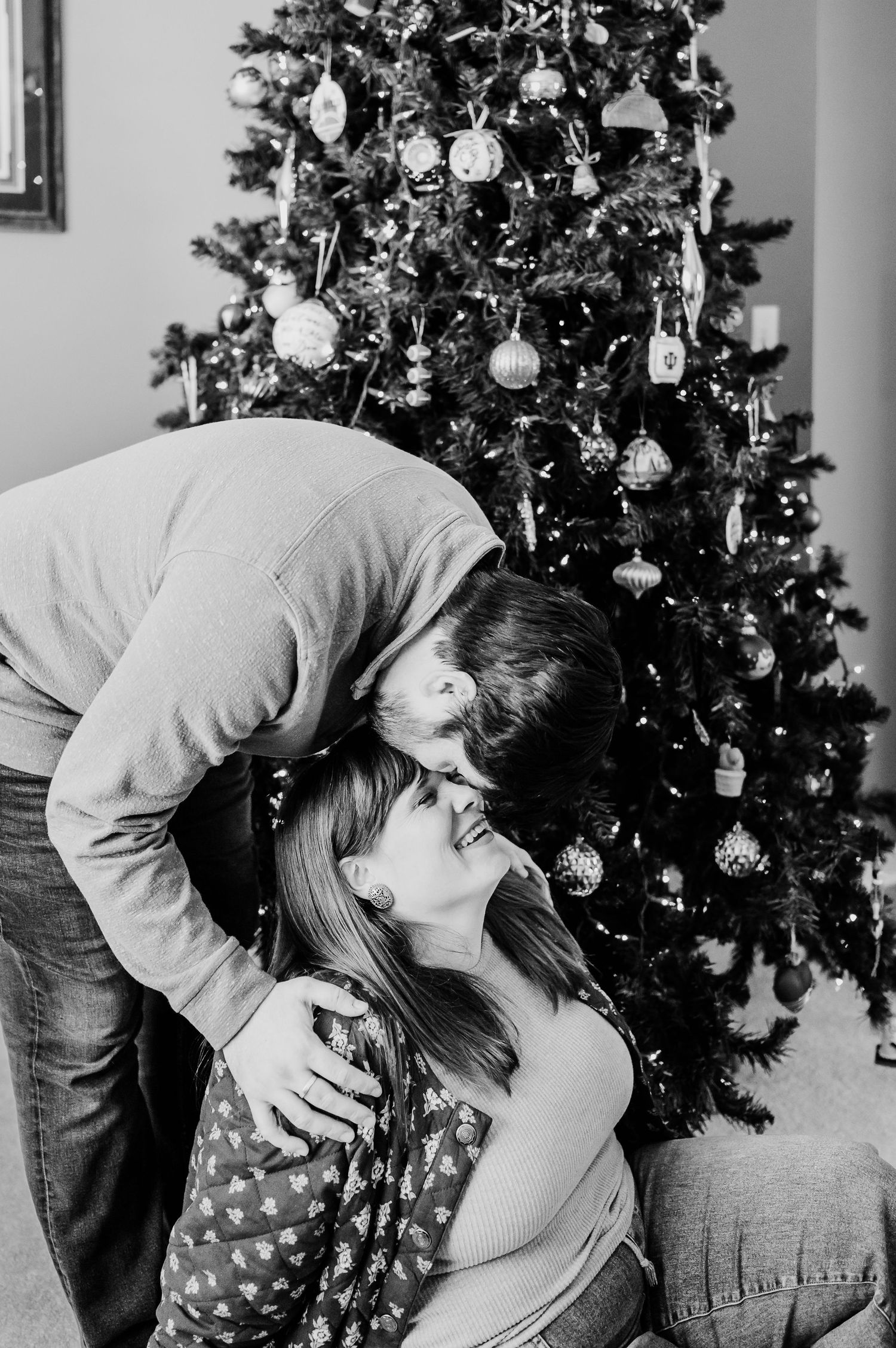 Black and white photo of a couple sharing a romantic kiss in front of a decorated Christmas tree indoors.