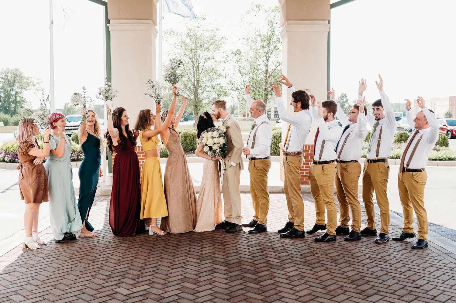 Wedding party in earth tones and suspenders poses together under covered outdoor archway.