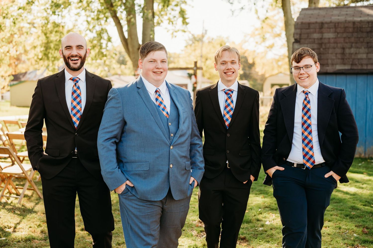 A group of four groomsmen stand together outdoors wearing suits and ties on a sunny autumn day.