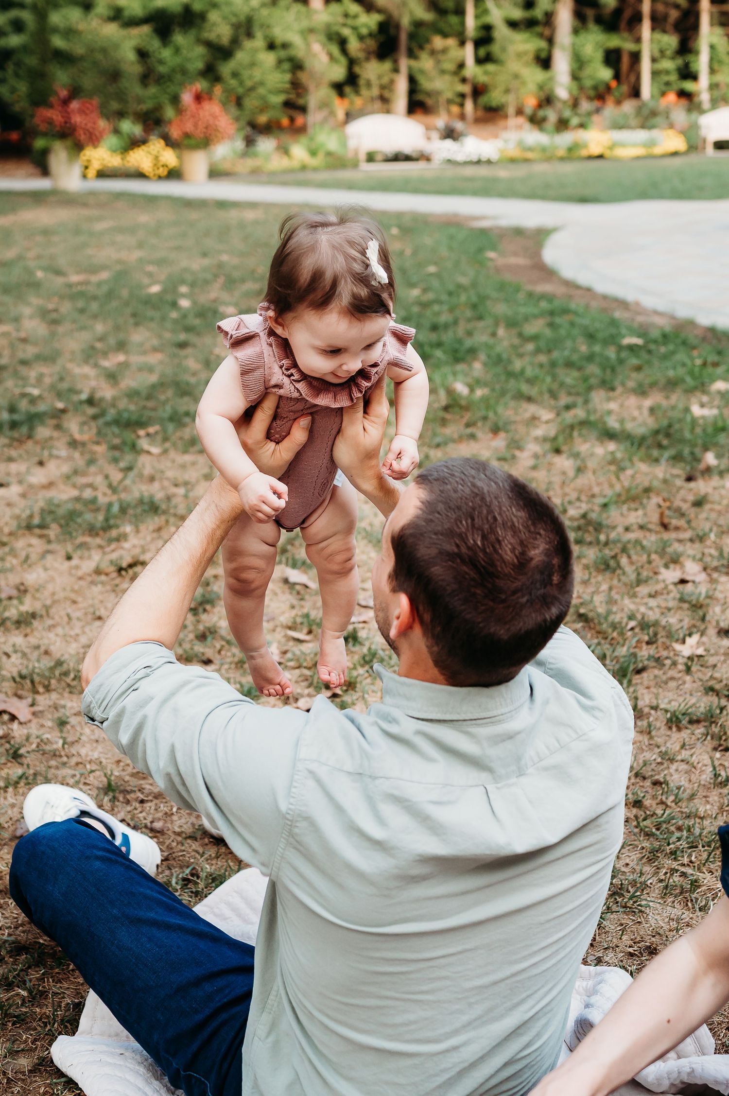Dad playing and lifting baby girl outdoors on grassy area during a sunny day at Newfields in Indianapolis.