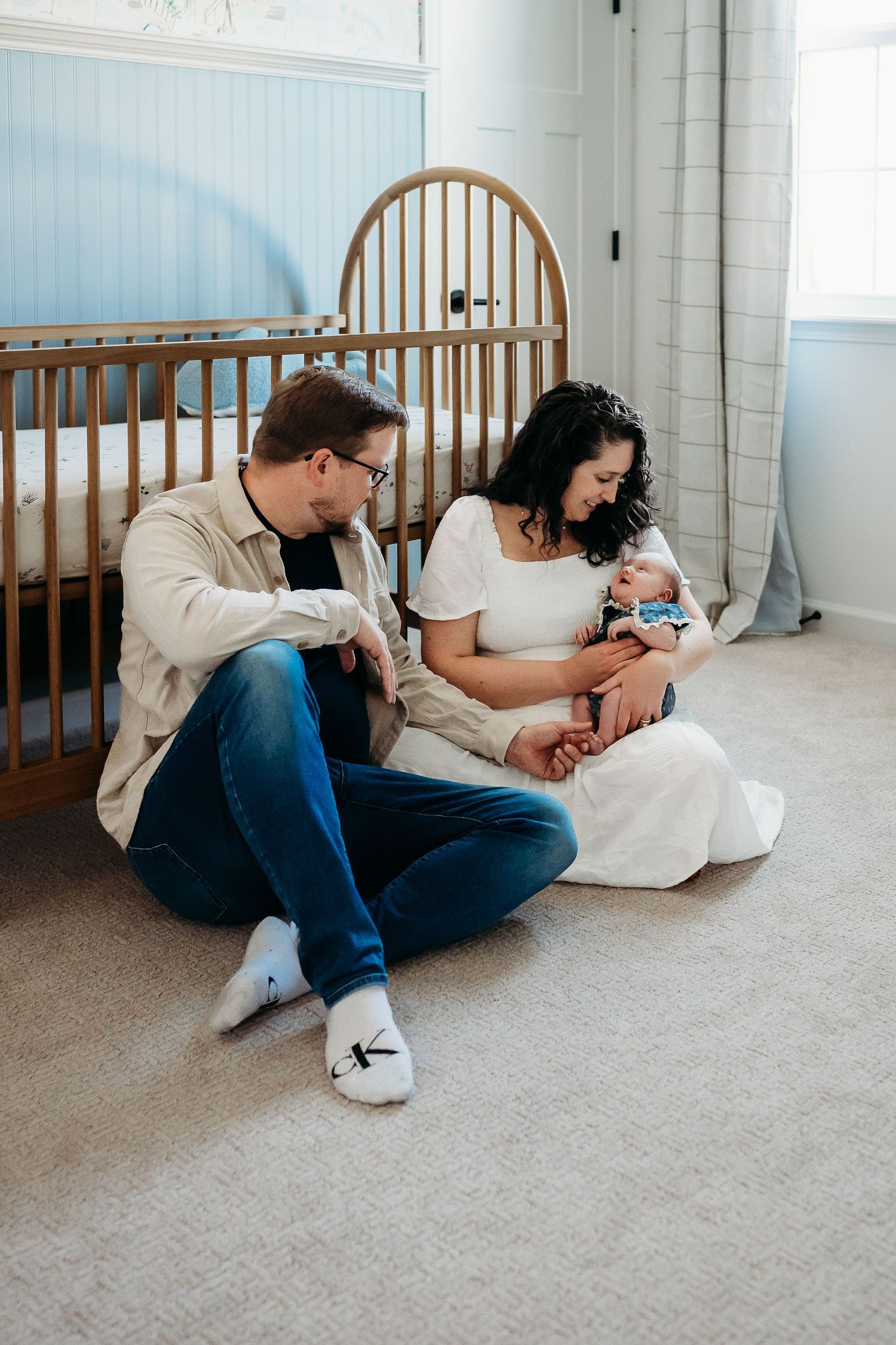 Parents sitting on nursery floor with newborn baby, wooden crib and blue walls in background.