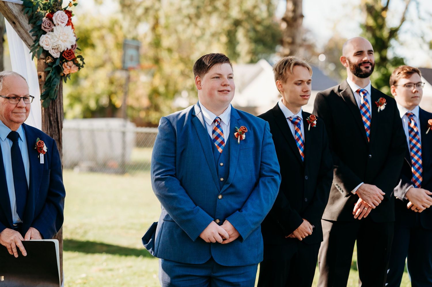 Wedding party members in blue suits and formal attire stand in line during outdoor autumn ceremony.