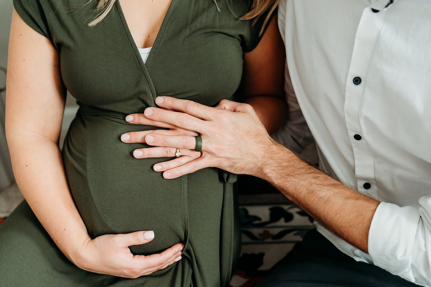 A pregnant person in green dress holds their belly while a partner's hand rests lovingly on top during a tender moment.