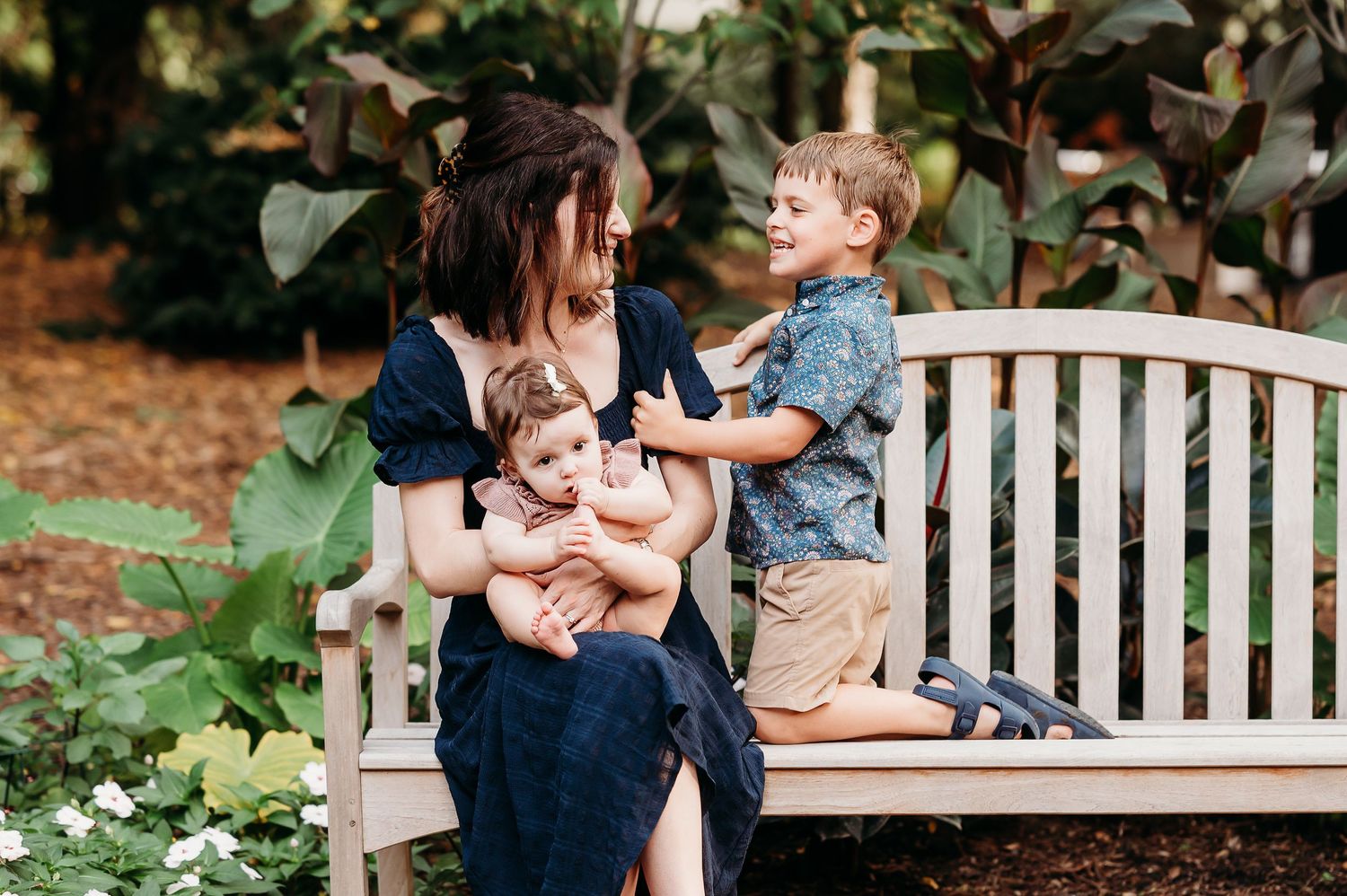 Sweet moment of mom with son and daughter captured on a white garden bench surrounded by lush greenery at Newfields in Indianapolis.