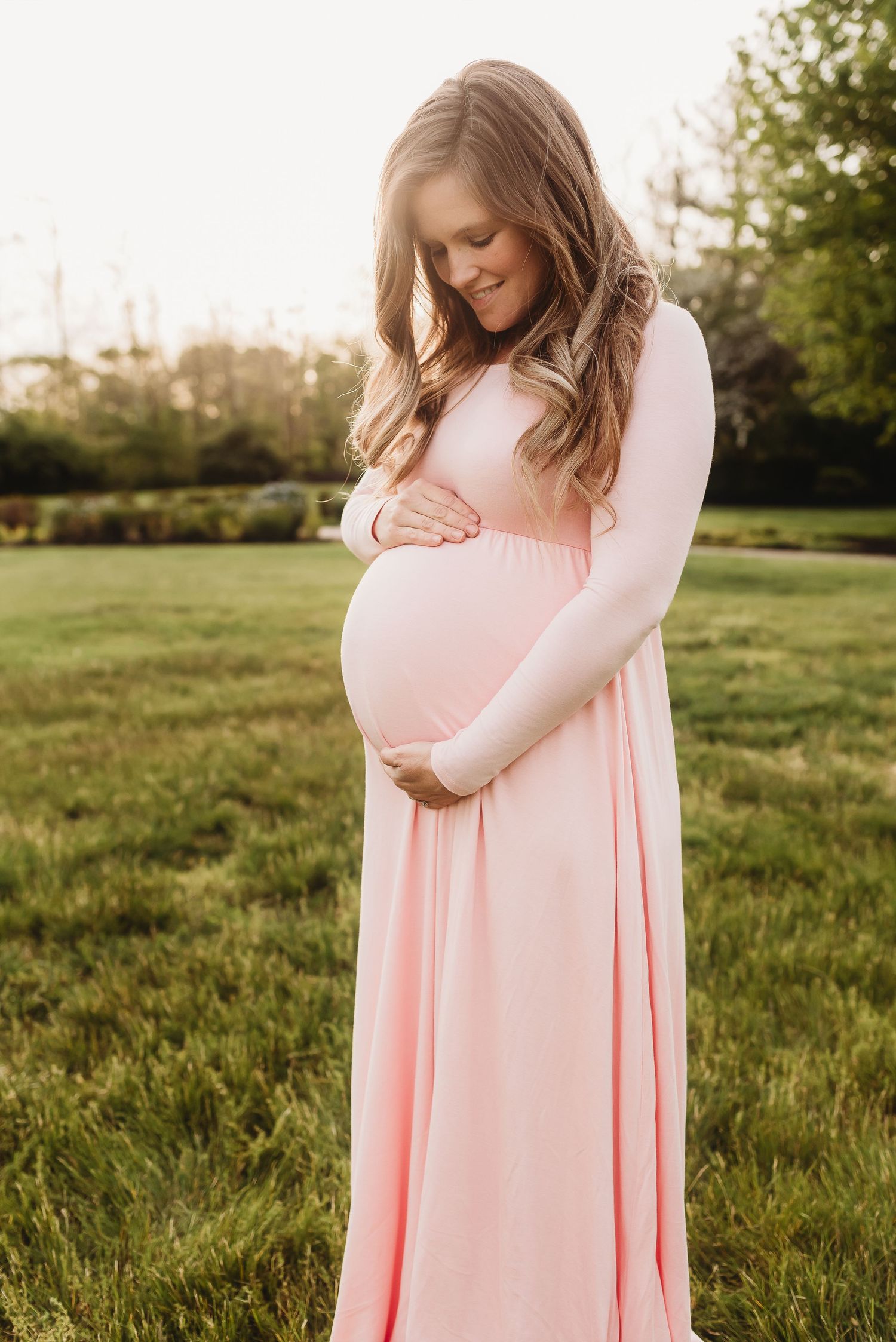A pregnant woman in a flowing pink dress cradles her belly while standing in a sunlit grassy field.