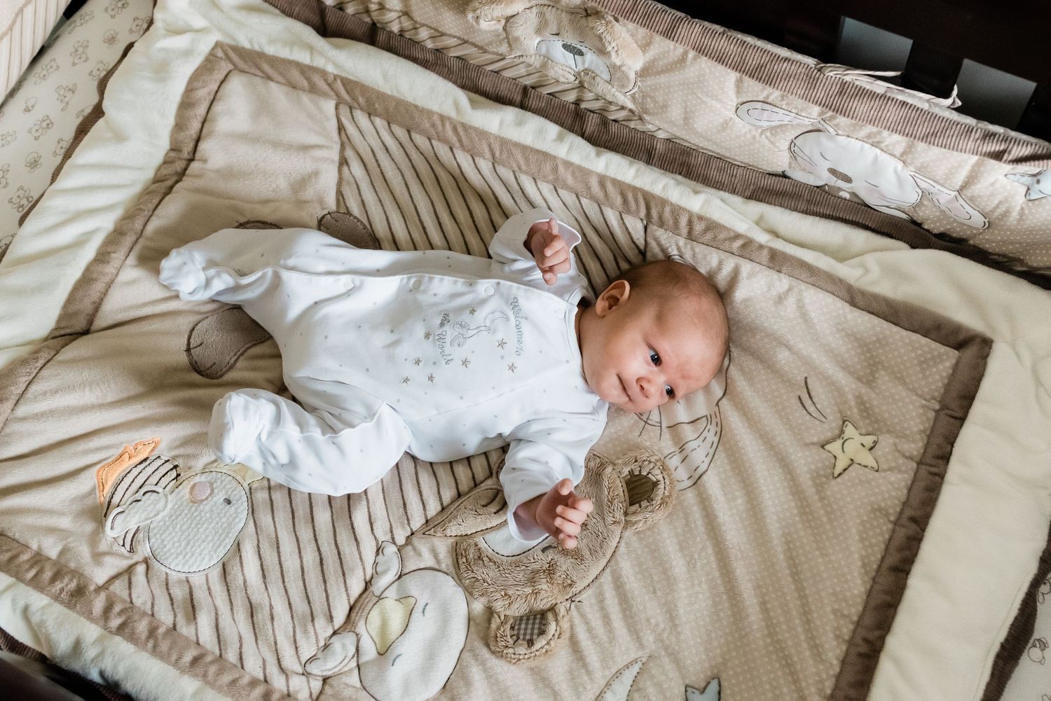 Baby in white onesie lying on patterned beige nursery bedding.
