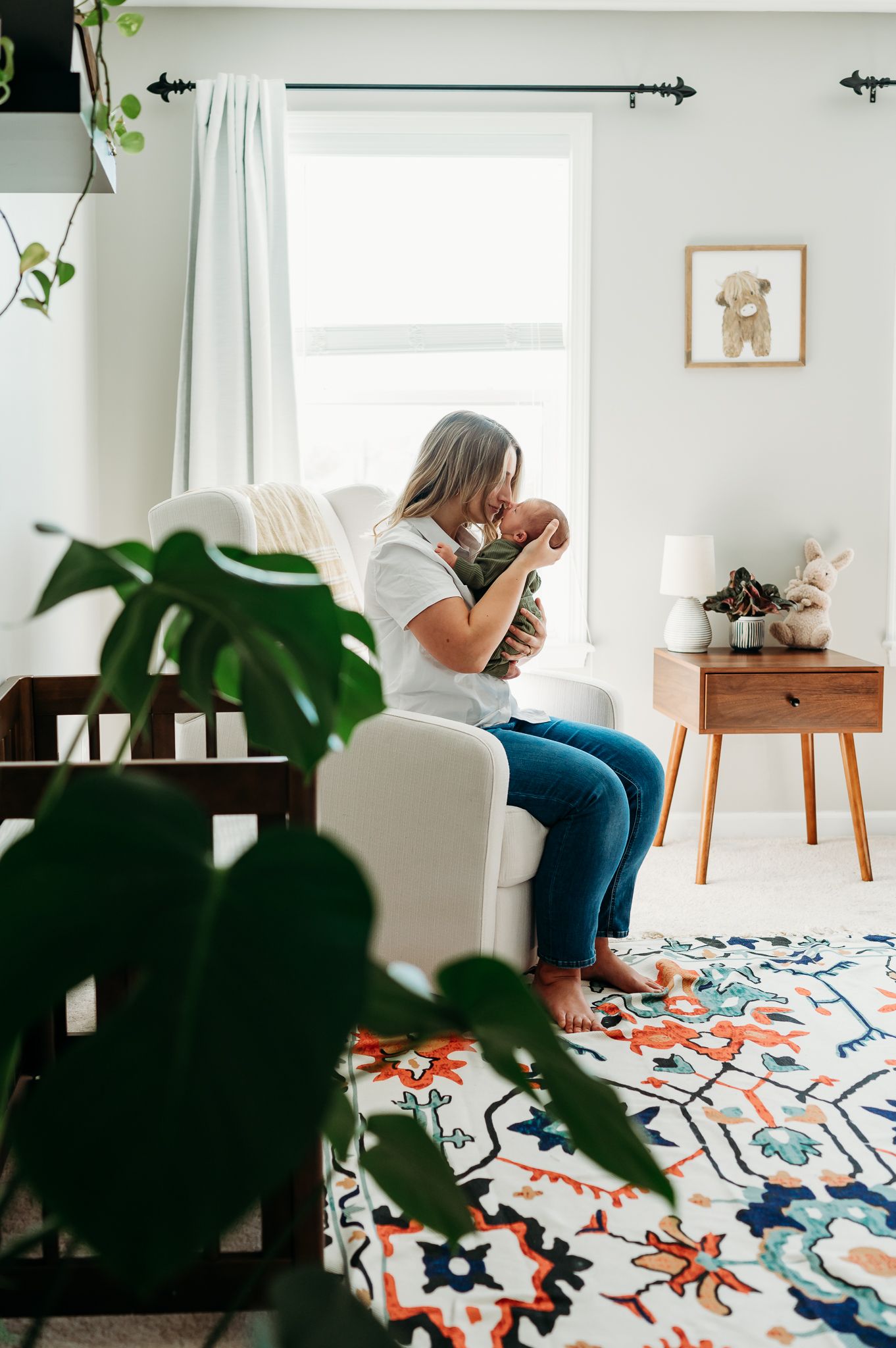 Mother kisses baby boy in cozy nursery featuring white curtains, a colorful patterned rug, potted plants and mid-century modern side table.
