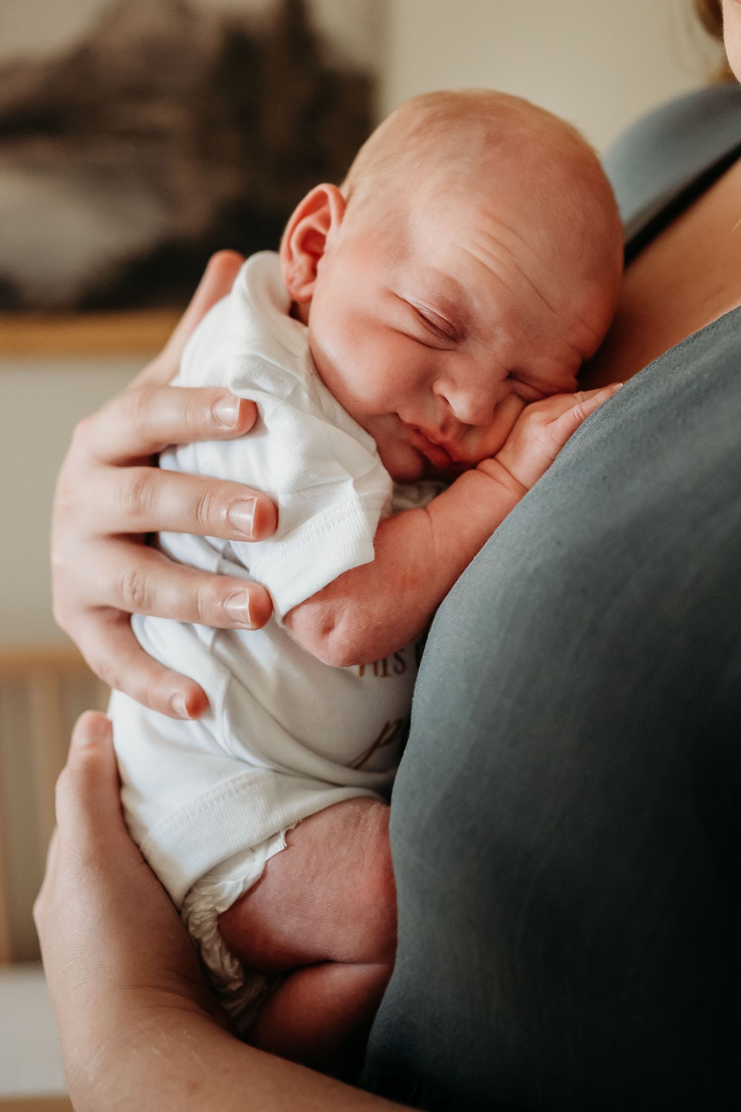 A newborn baby sleeping peacefully in an adult's arms, dressed in a white onesie.