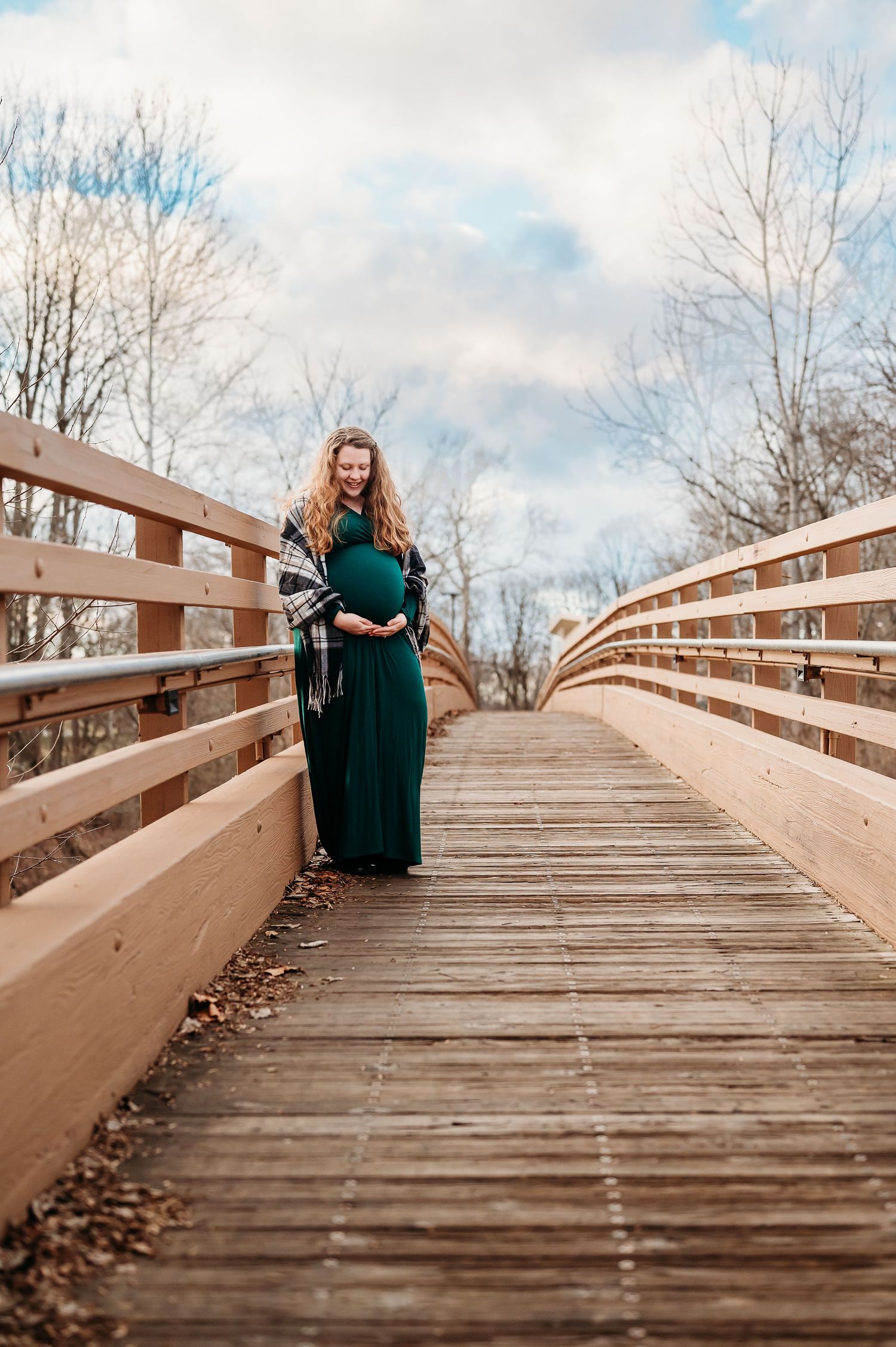 Pregnant woman in green dress on wooden bridge, smiling and cradling belly, surrounded by bare trees.